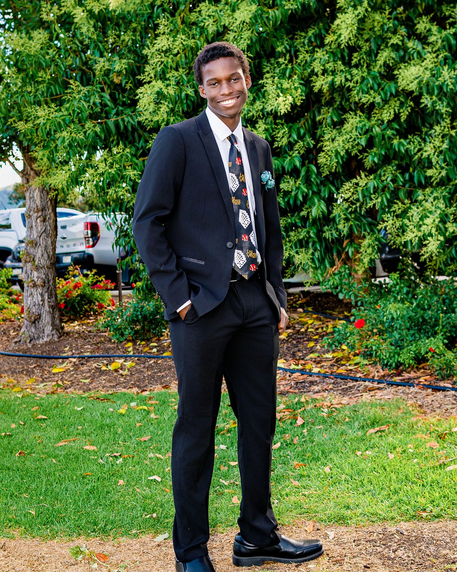 Young man in black suit and patterned tie smiling outdoors in a garden setting.