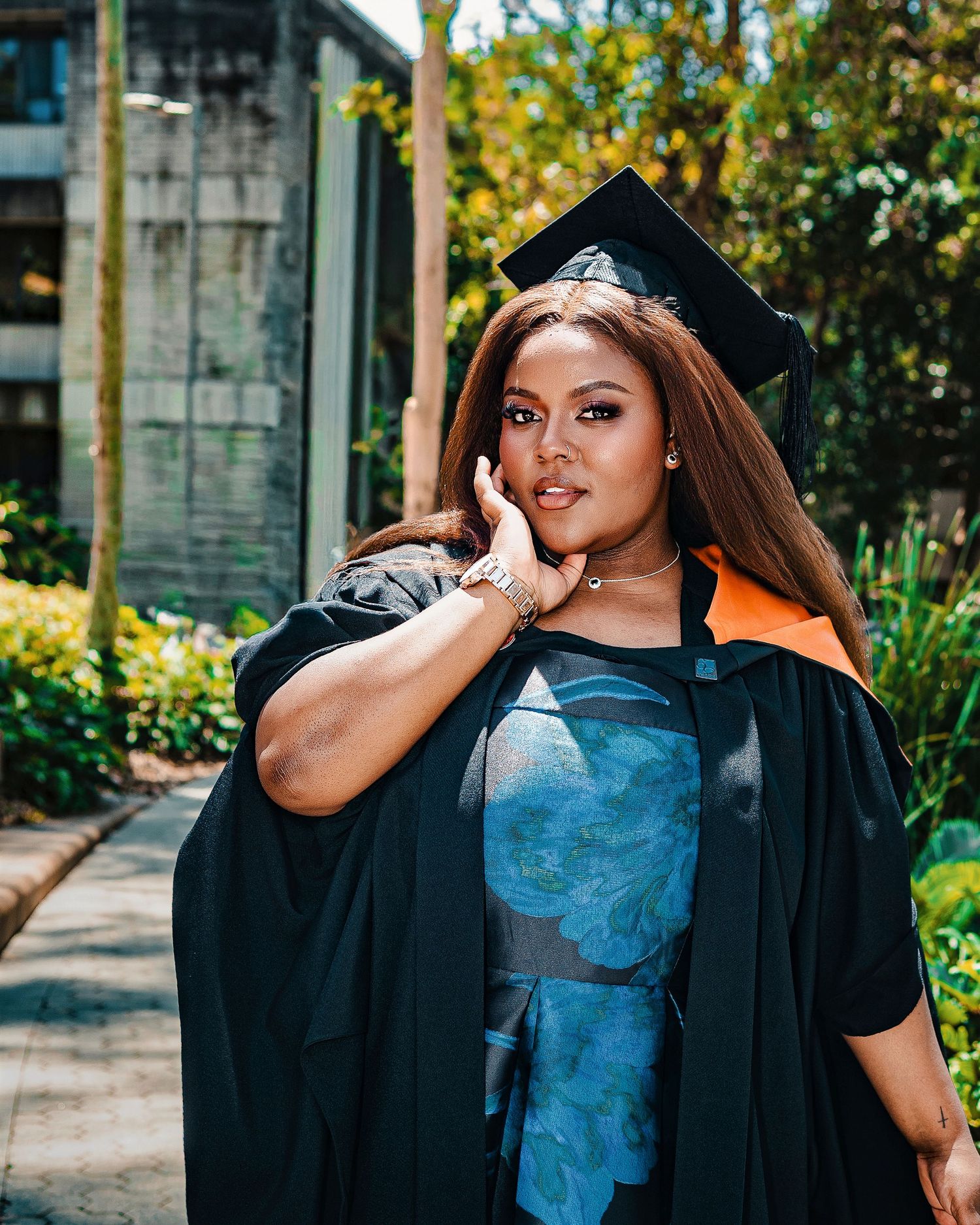 A graduate in black cap and gown with orange trim poses proudly in a lush outdoor setting.