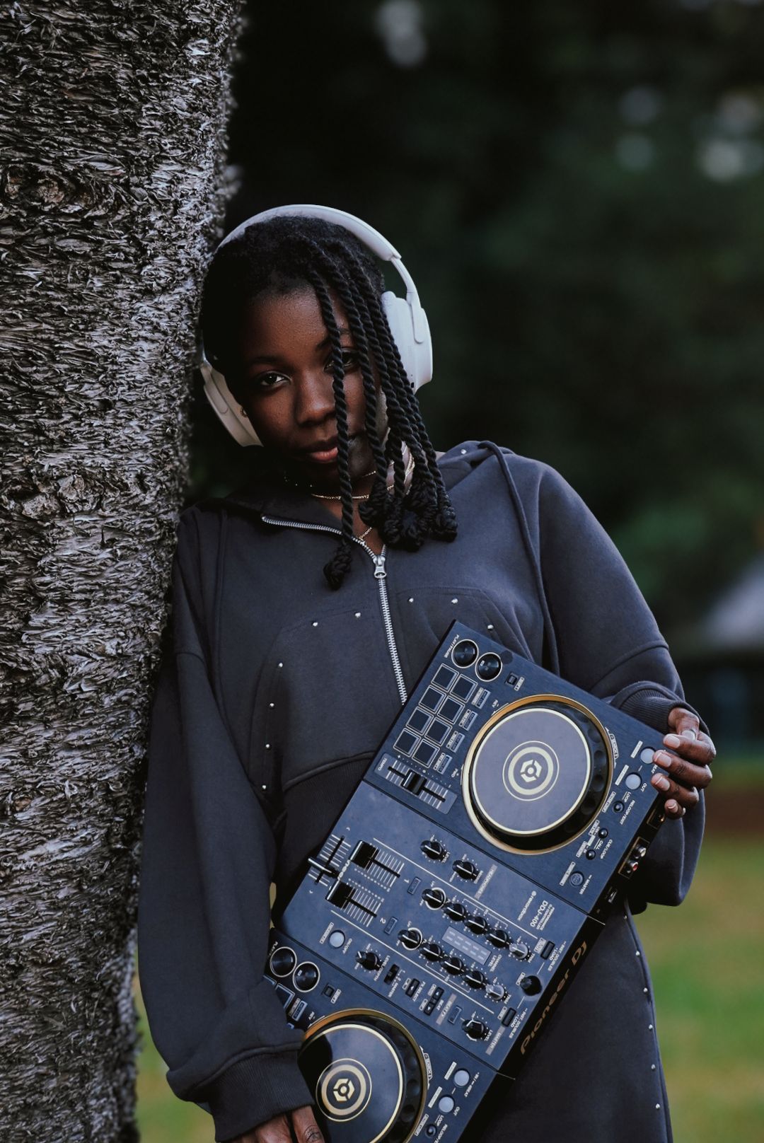 A young woman leans against a tree holding a DJ controller while wearing white headphones outdoors.