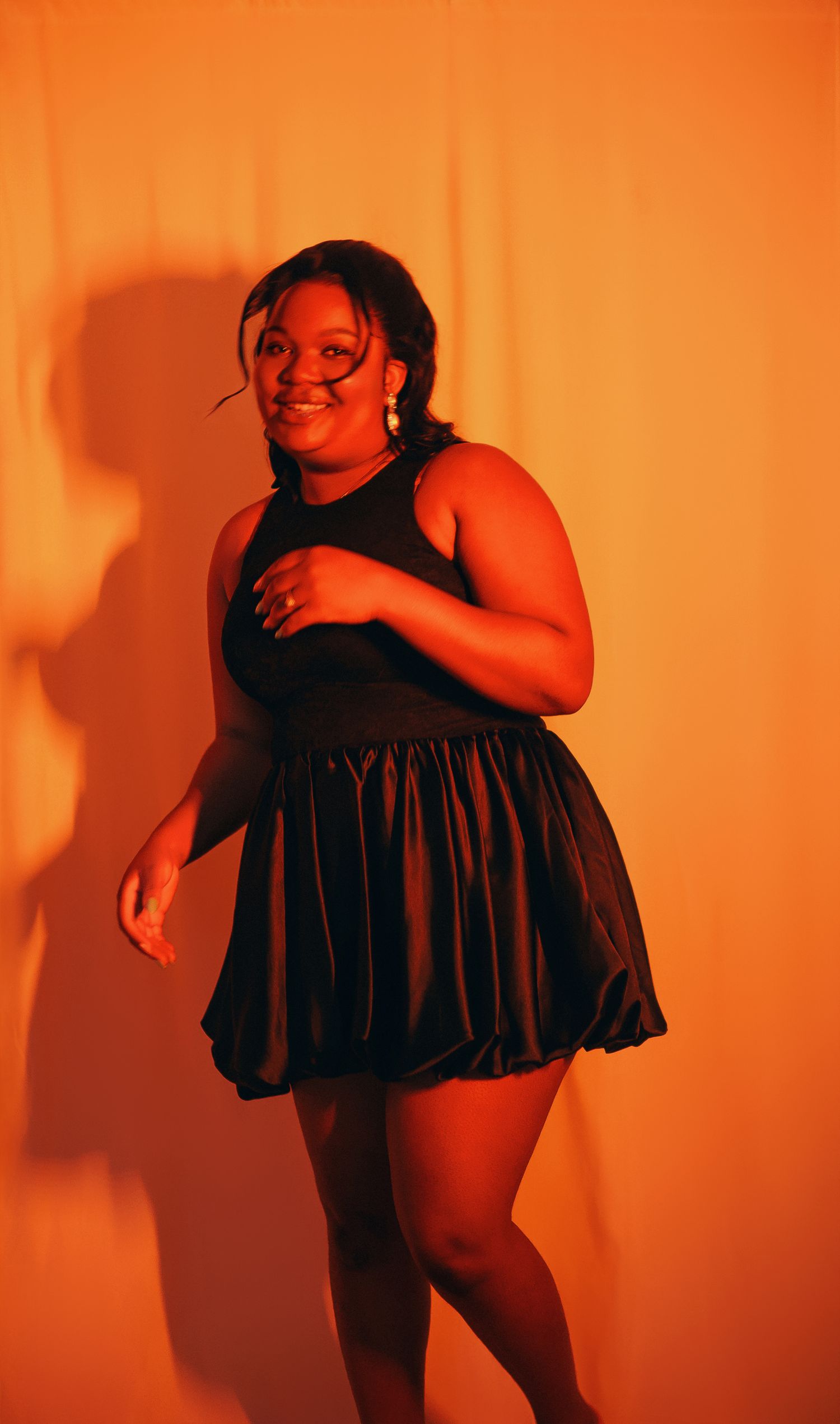 Smiling woman in black bubble skirt dress posing in orange lit studio.