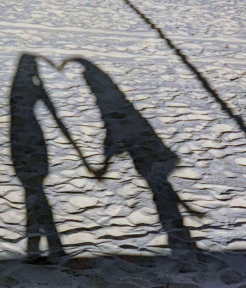 Shadows of two people forming a heart shape with their hands on sandy beach.