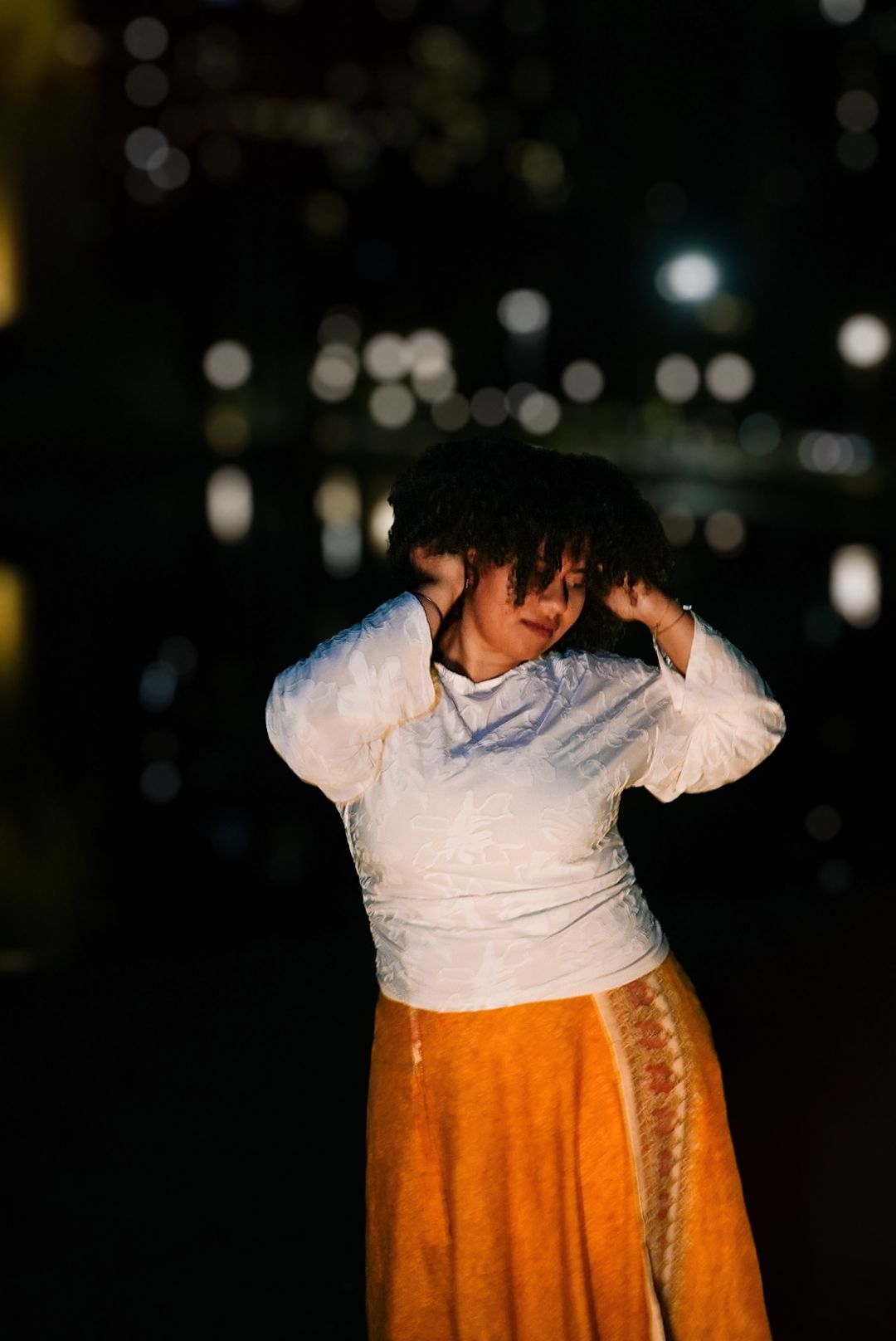 Woman in white top and orange skirt adjusting her curly hair during a nighttime outdoor photo shoot.