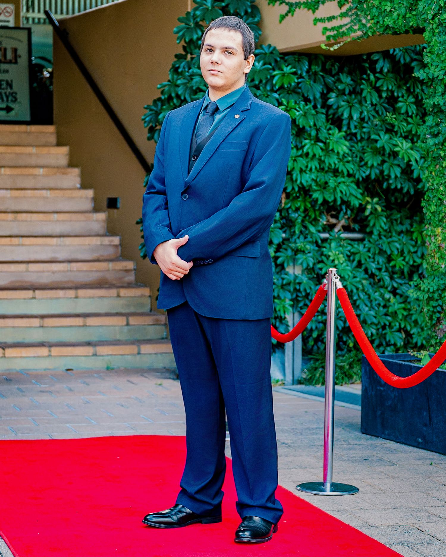 Man in navy blue suit standing on red carpet at formal event entrance with rope barriers.