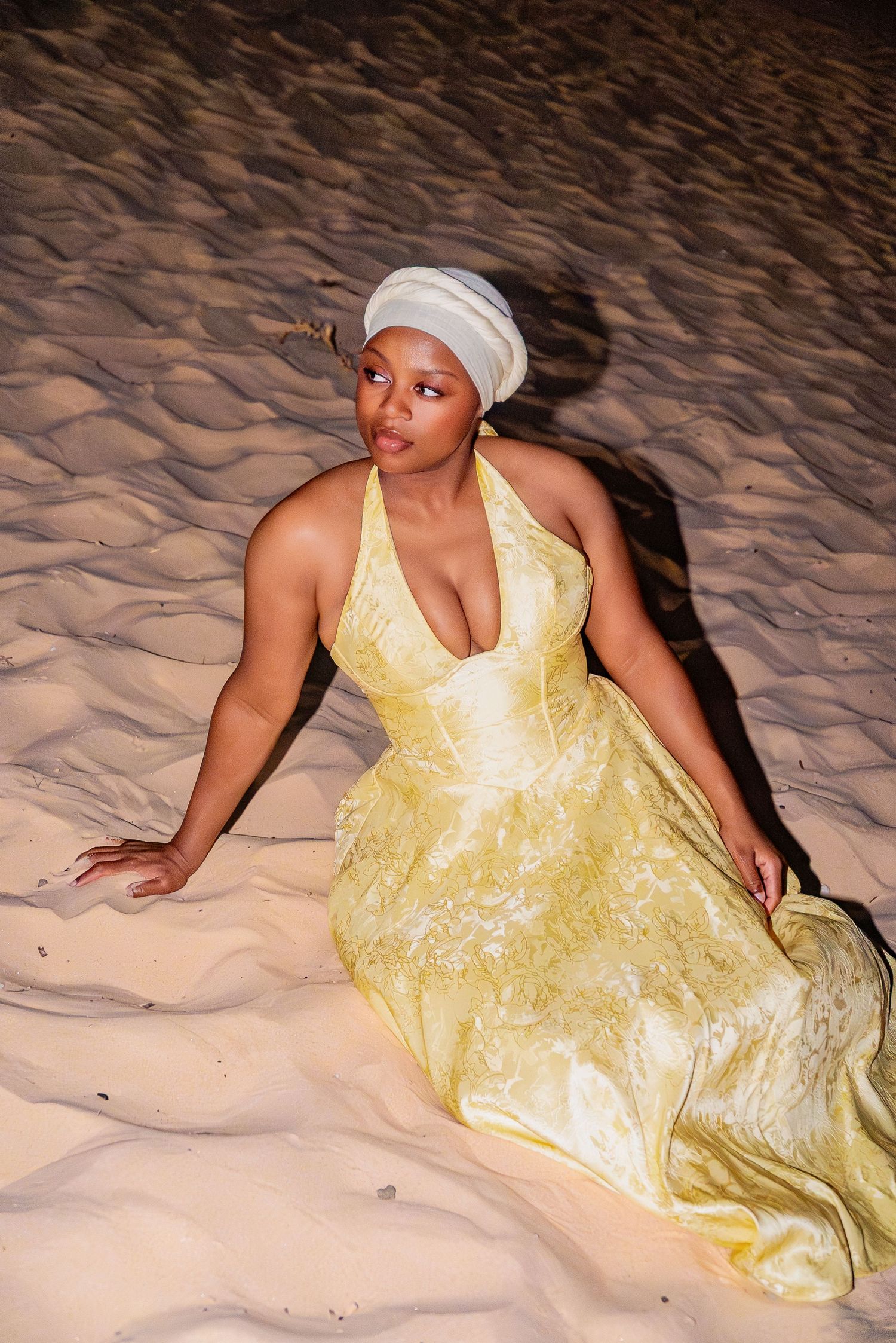A woman in a yellow halter gown and white turban reclines gracefully on rippled sand dunes at night.