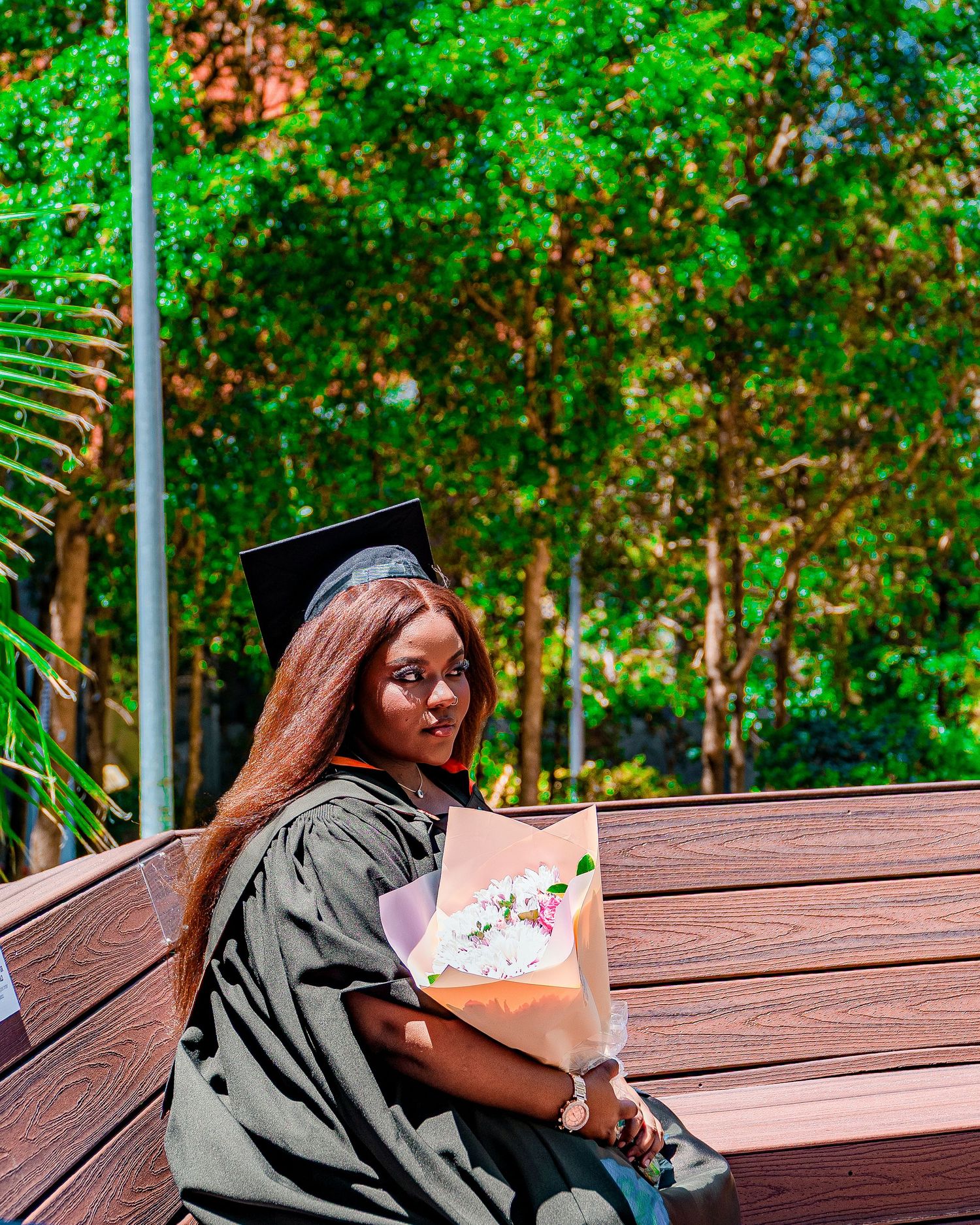 Graduate in cap and gown sits on bench holding bouquet of flowers outdoors.