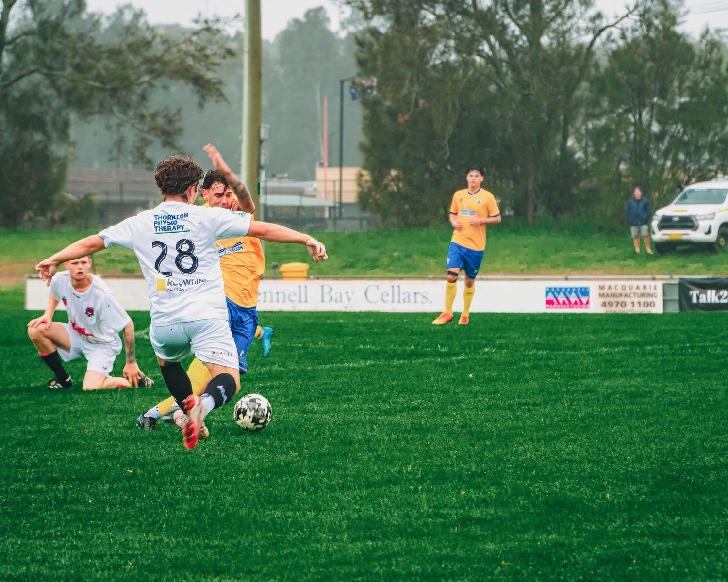 Youth soccer players compete on a green field, with player number 28 dribbling the ball during a match.