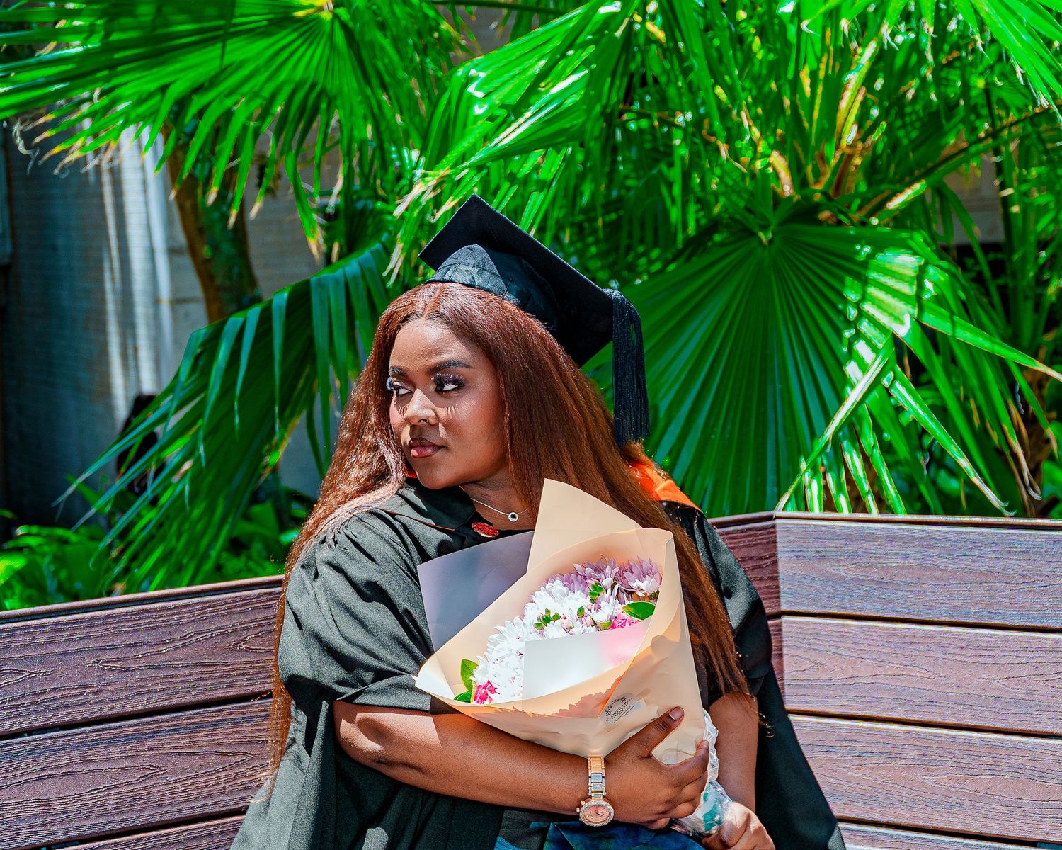 Graduate in black cap and gown holds colorful flower bouquet while sitting on wooden bench outdoors.