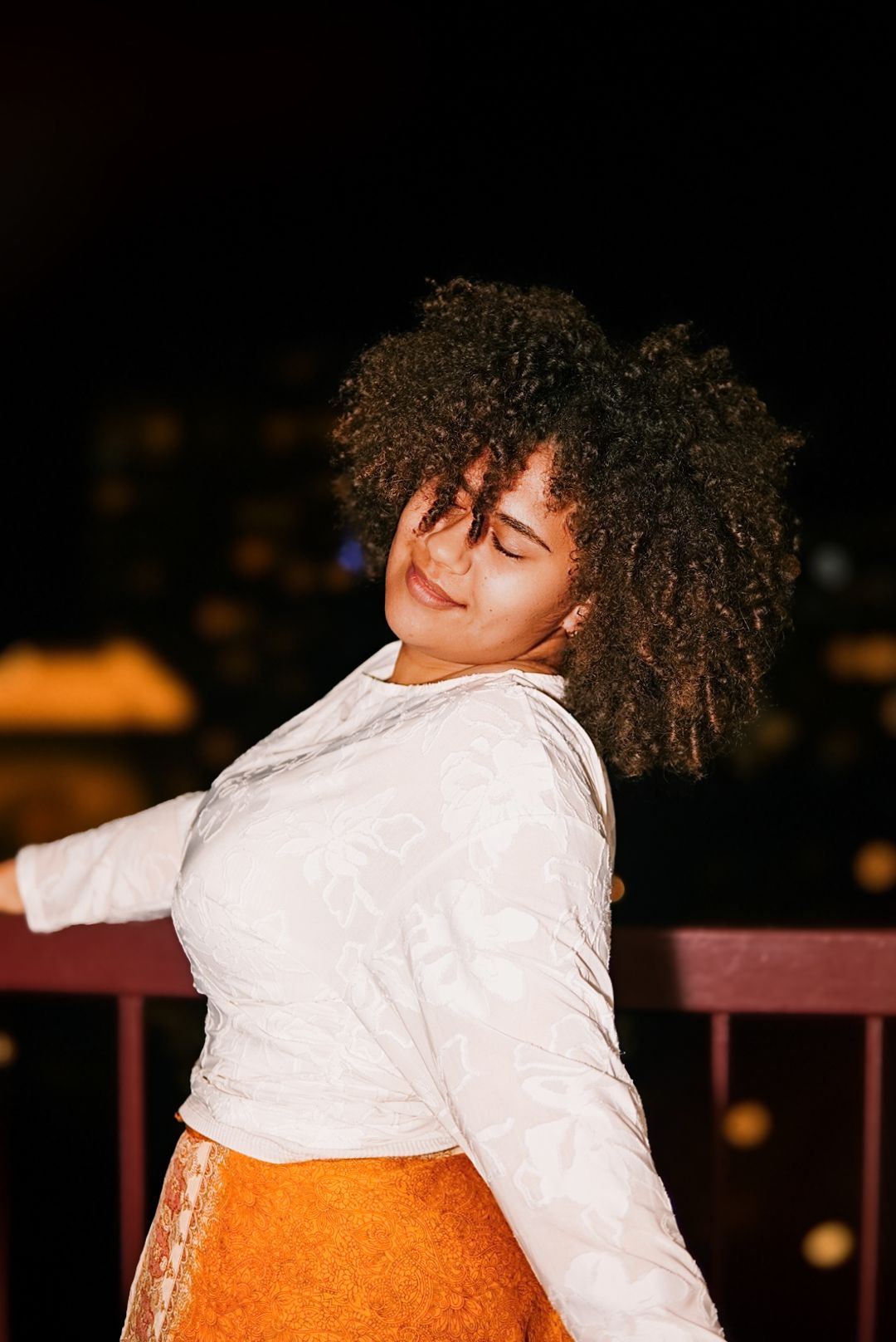 Woman with curly afro hair in white top and orange skirt spinning joyfully during a night photoshoot.