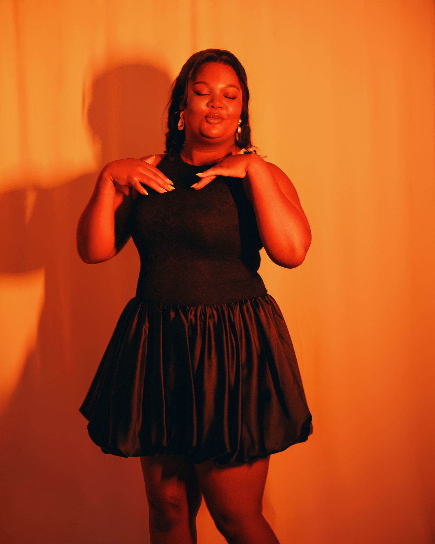 A plus-size woman poses confidently in a black bubble mini dress against a warm orange-lit background.