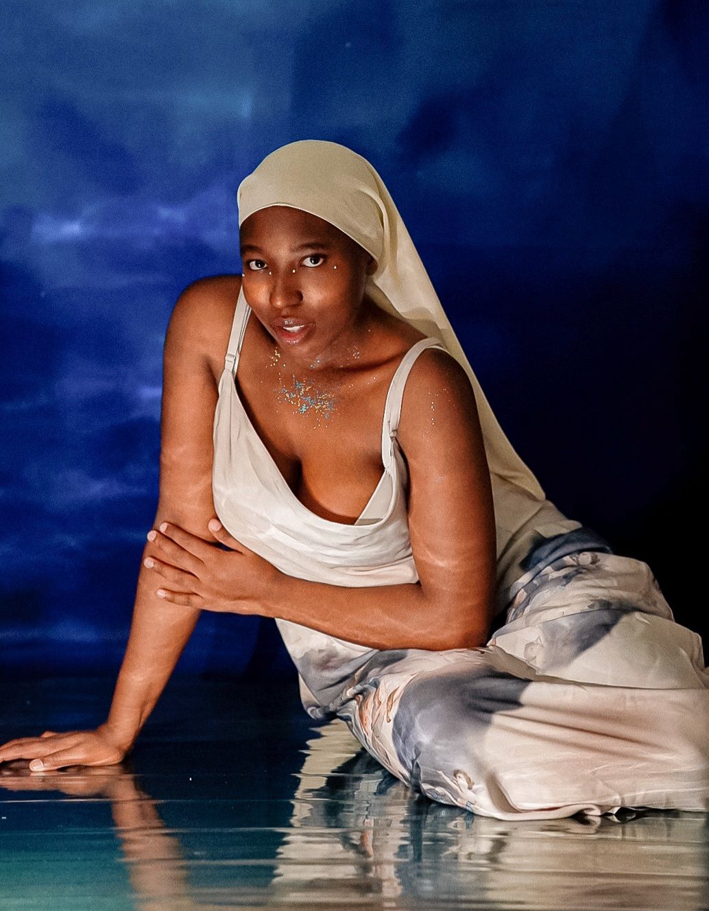 A woman in white draped fabric and headscarf poses gracefully against a dramatic blue backdrop.
