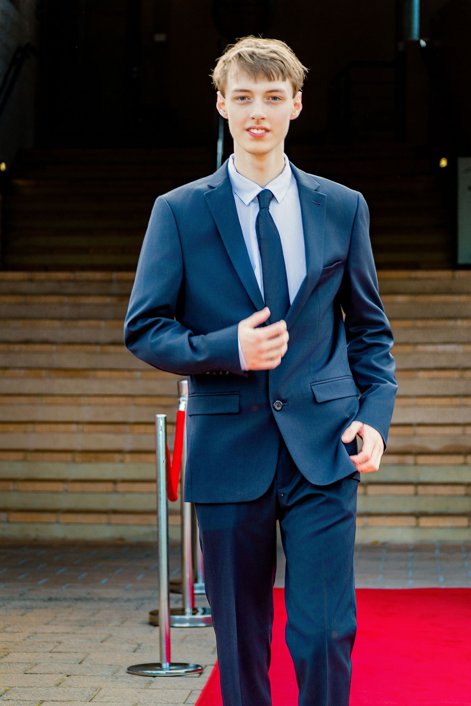 Young man in navy blue suit and tie walking on red carpet, smiling confidently at formal event.