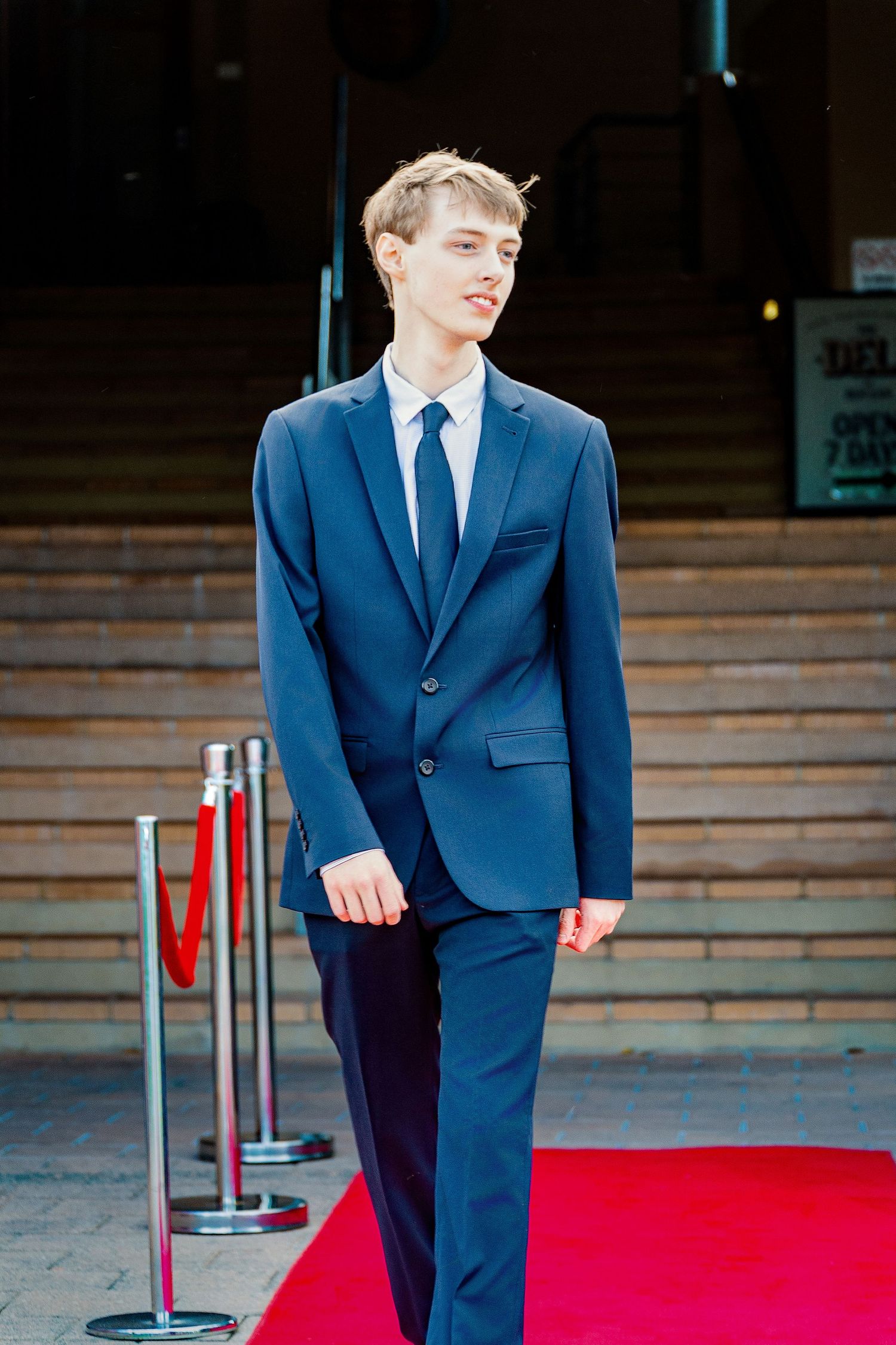 Young man in navy blue suit and tie walking on red carpet outside formal venue.