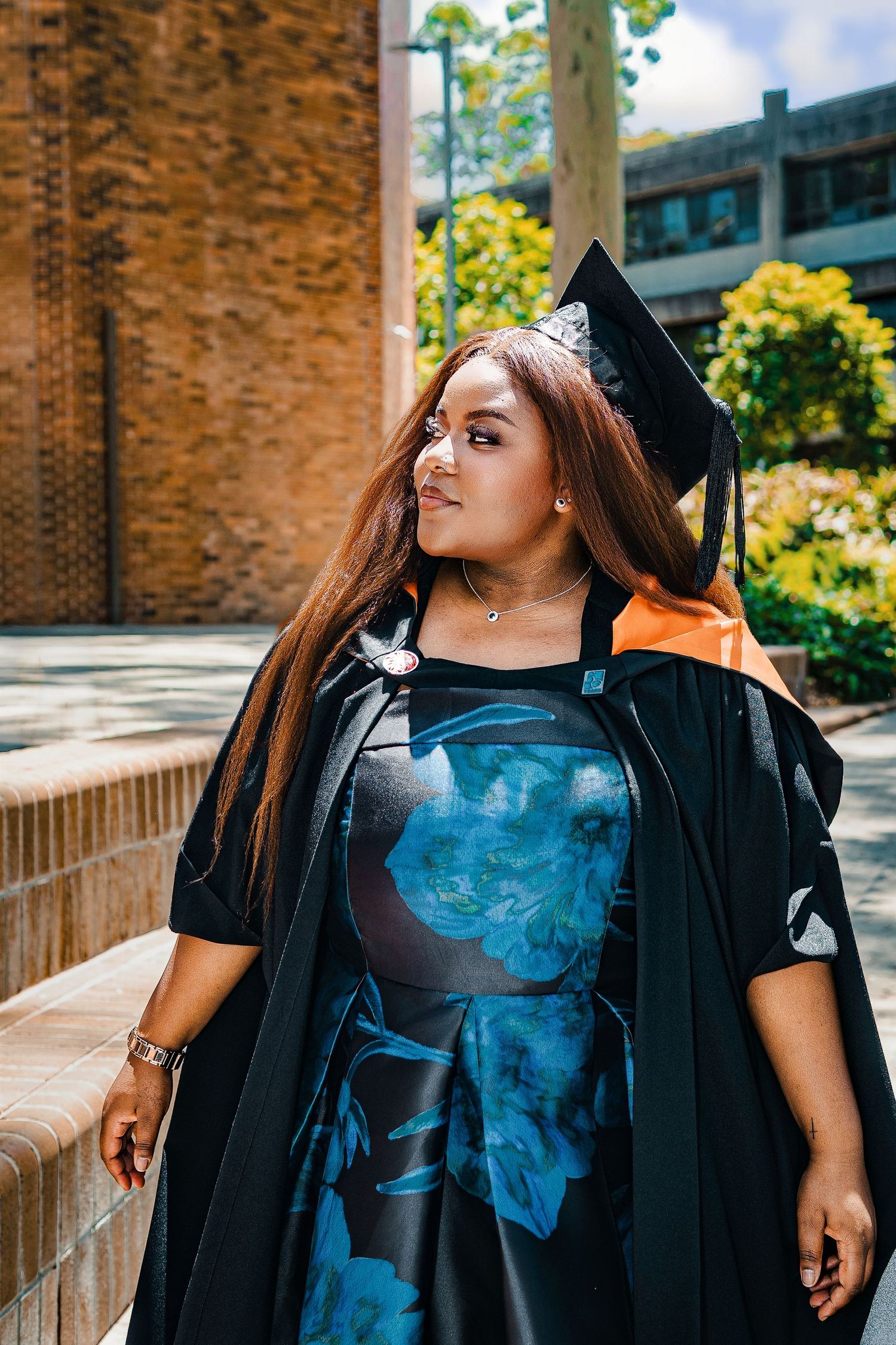 Woman in black graduation gown and cap with orange trim poses outdoors near brick building.