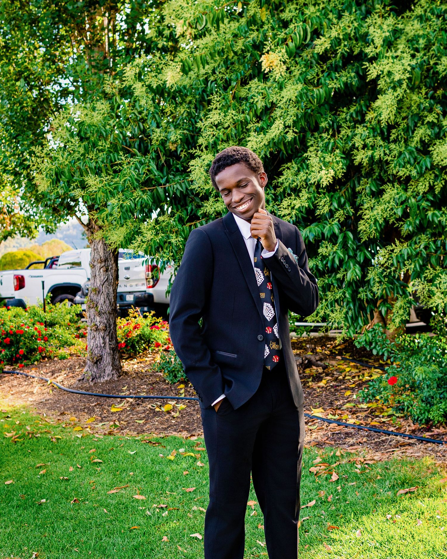 Young man in navy suit and patterned tie smiling outdoors in garden setting.