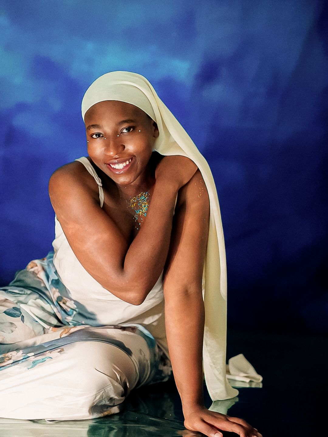 Smiling woman in white hijab and floral dress poses gracefully against a deep blue backdrop.