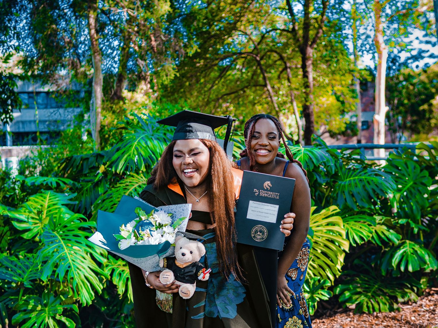 Two women posing outdoors at graduation, one wearing cap and gown holding flowers and teddy bear, other holding diploma.