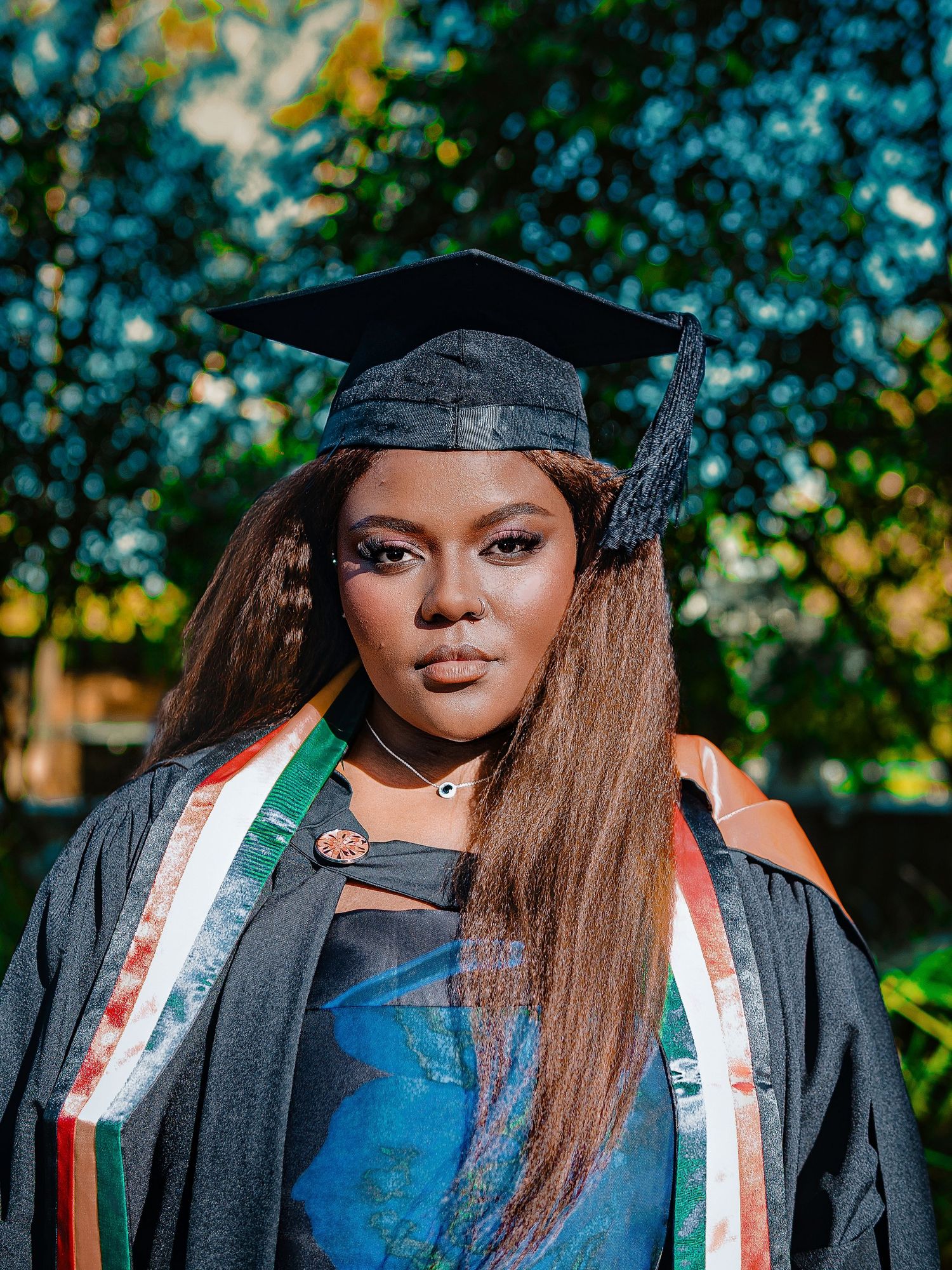 Young female graduate in black mortarboard and gown with colorful stole poses among green trees.