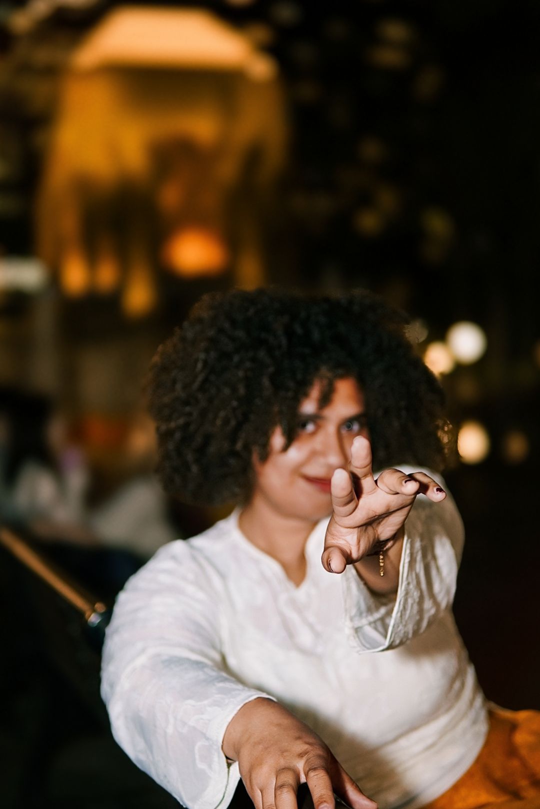 Curly-haired woman in white top reaches toward camera with playful expression at night.