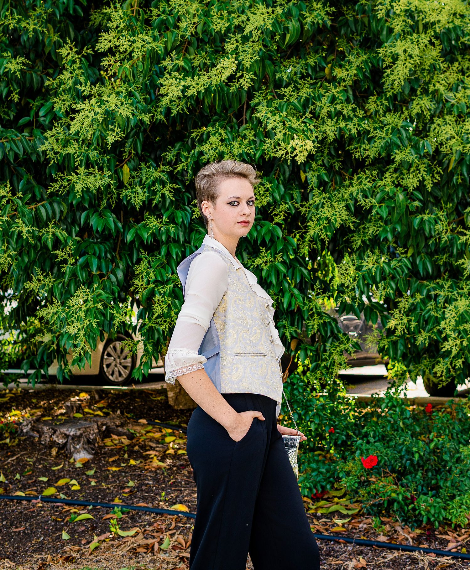 Young woman with short hair poses in a garden wearing a white vest and black pants amid lush green foliage.