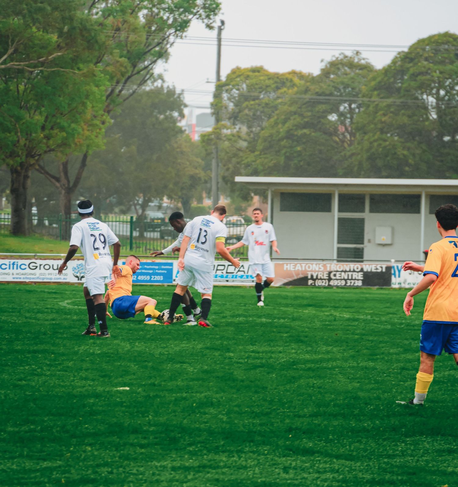Soccer players in white and yellow uniforms compete on a green field during an outdoor match on a cloudy day.