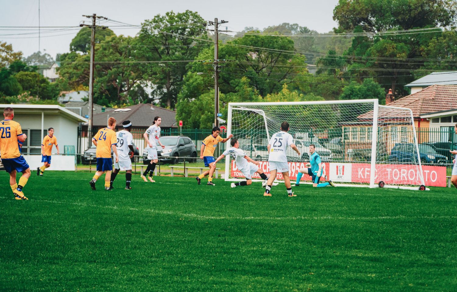 Soccer teams in yellow and white jerseys play near the goal during an outdoor match.