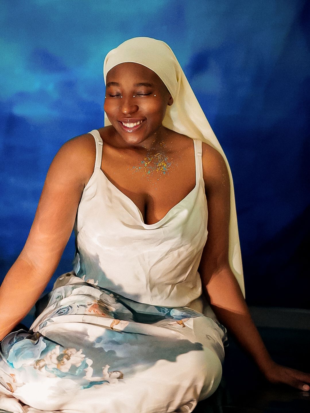 Smiling woman in white dress and head covering poses against blue background with glitter chest decoration.