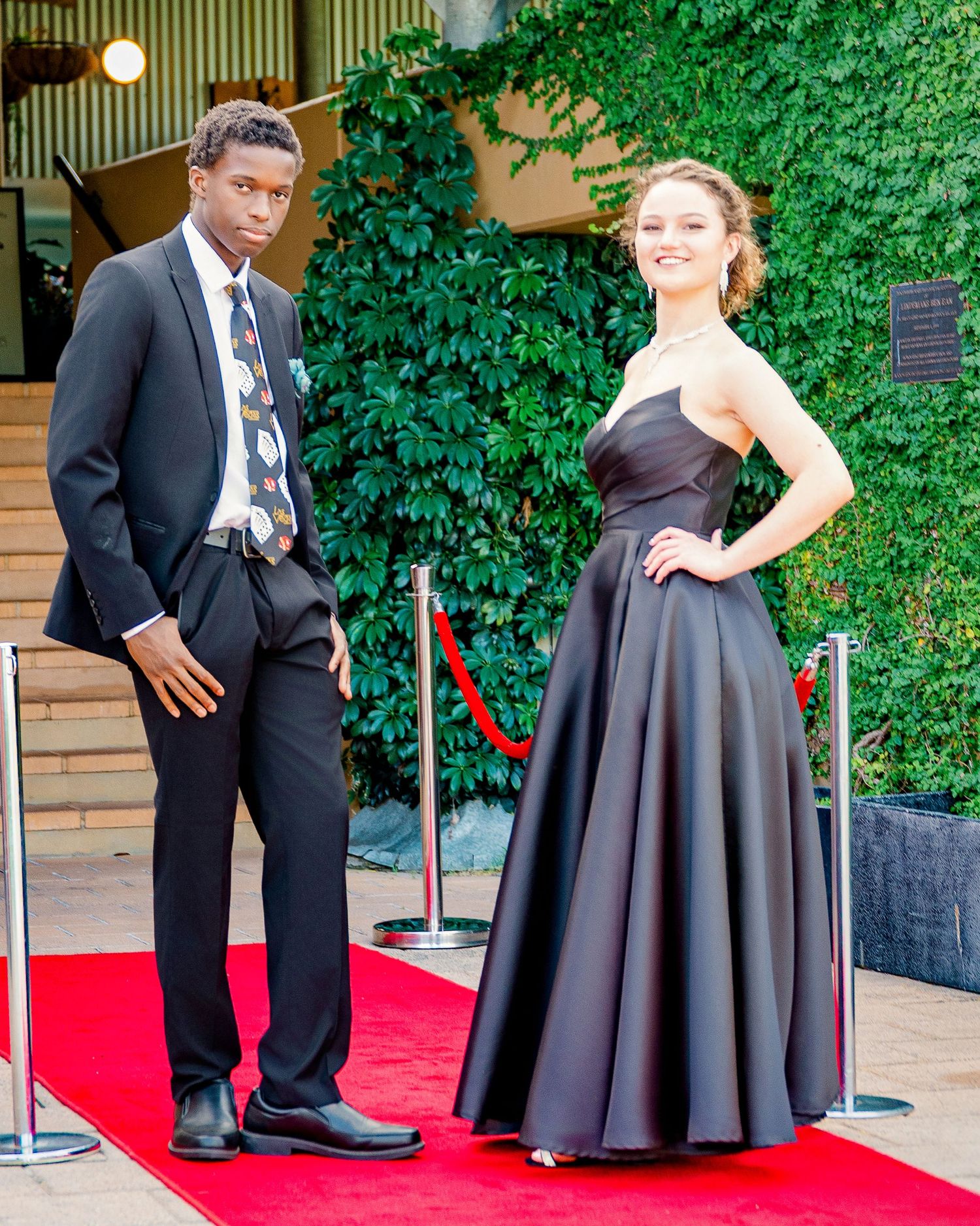 Young couple dressed formally pose together on red carpet at outdoor evening event.
