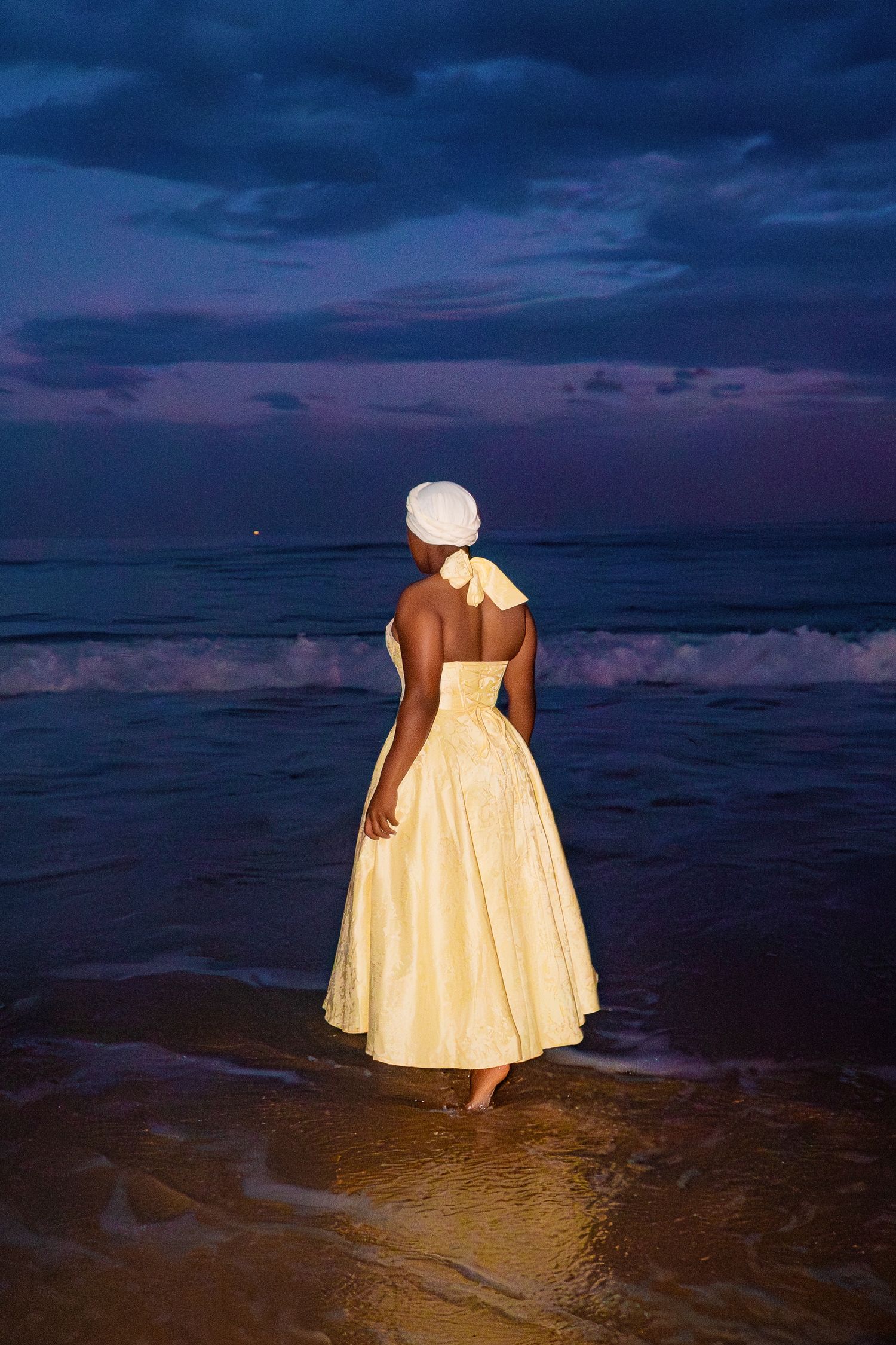 A woman in a yellow ball gown and white turban stands barefoot facing the ocean under a dramatic twilight sky.
