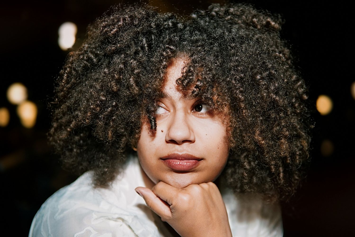 A young woman with natural curly hair rests her chin on her hand in a softly lit portrait.