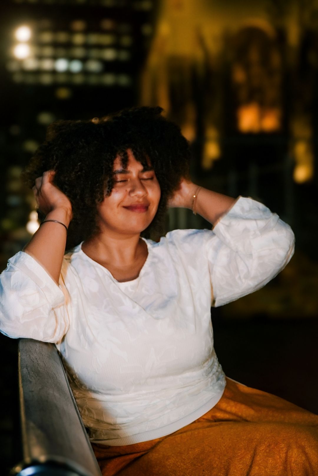 Woman with curly hair smiles with eyes closed, hands in hair, wearing white top at night.