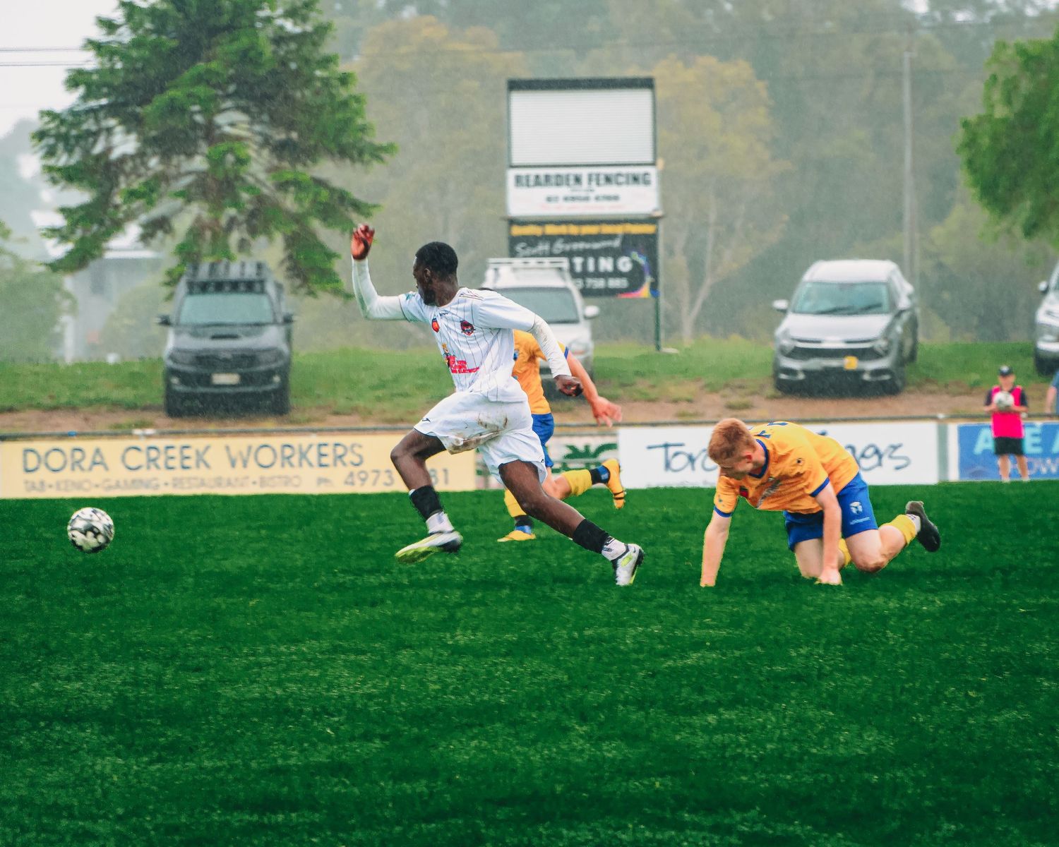 Soccer players compete on a green field, with one player dribbling past a sliding opponent during an outdoor match.