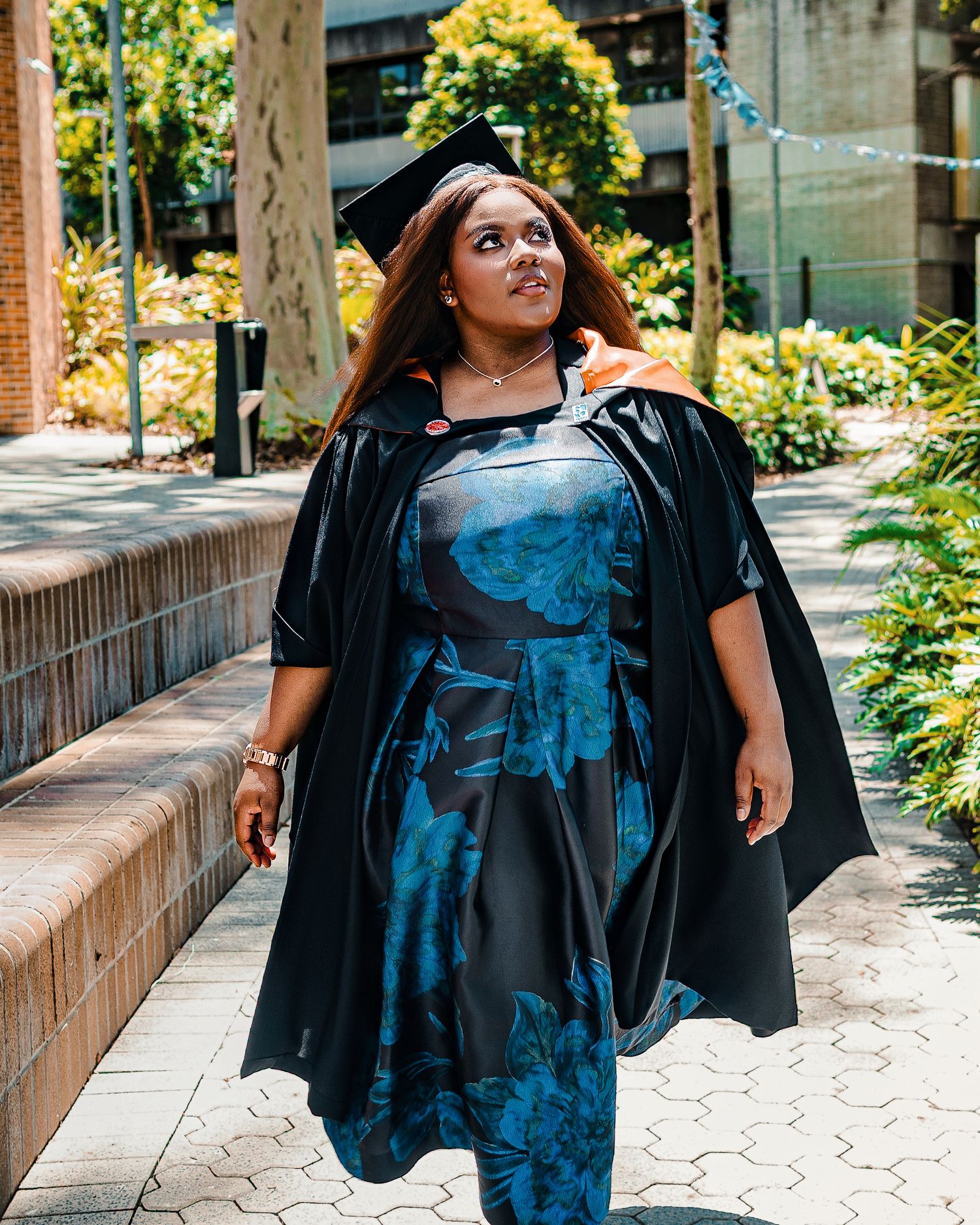 A young woman in graduation cap and gown over a blue floral dress walks along a brick pathway outdoors.