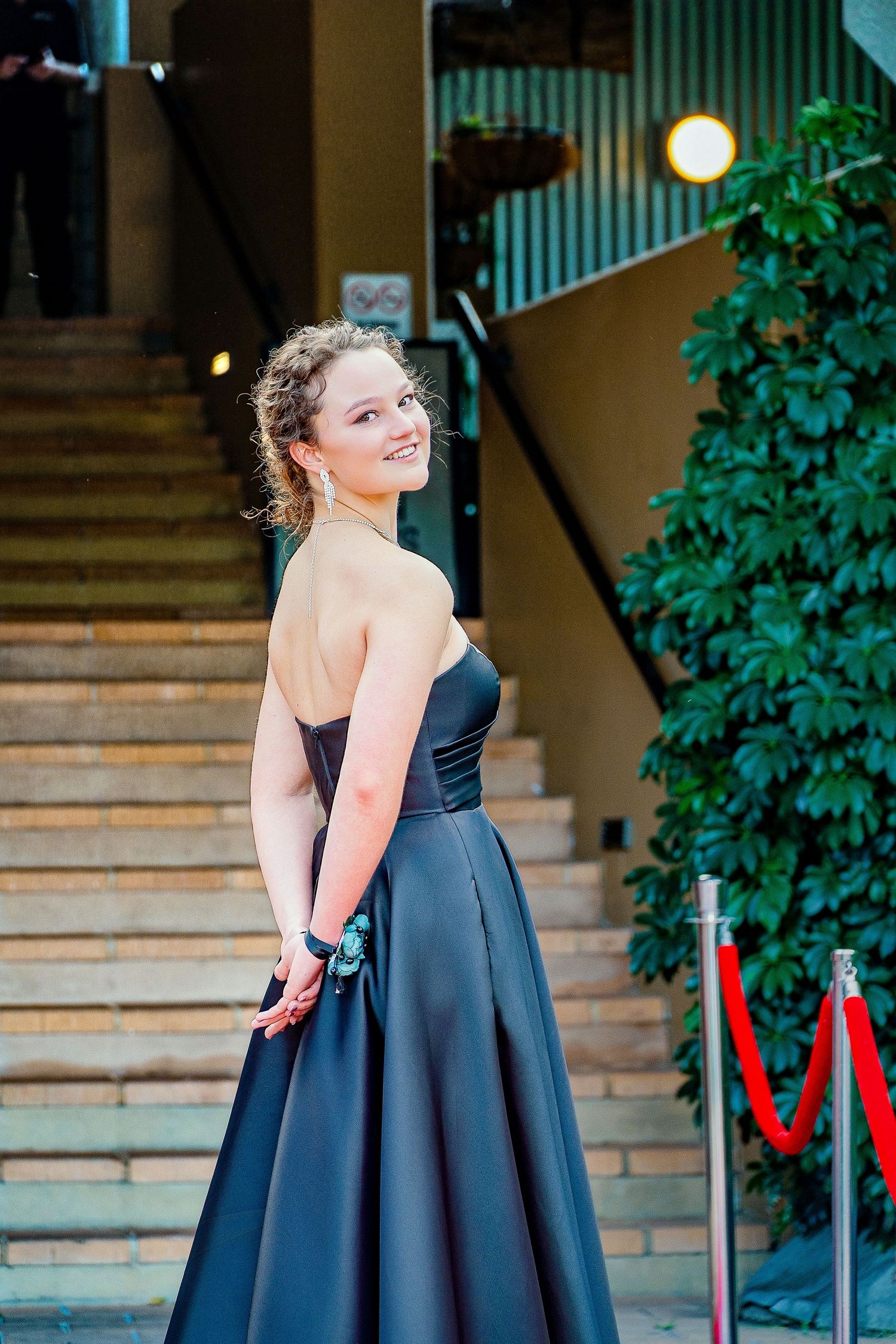 Young woman in elegant strapless black ball gown smiling over shoulder on red carpet event staircase.