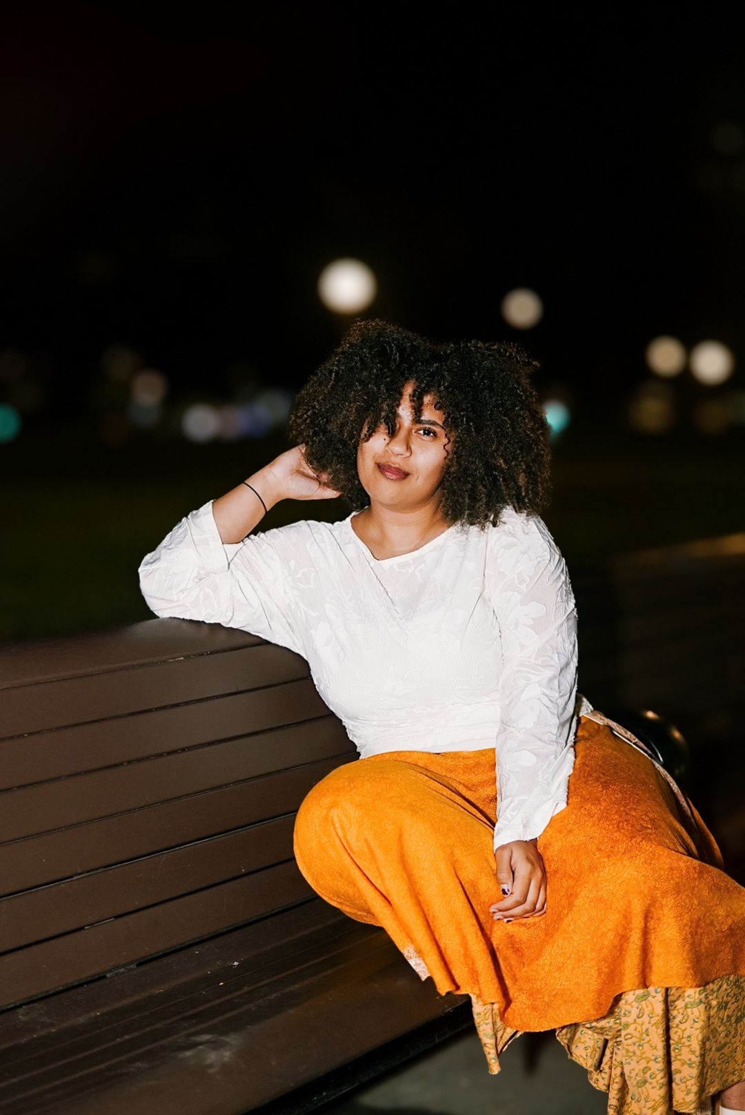 Woman with curly hair in white top and orange skirt sits relaxed on a park bench at night.