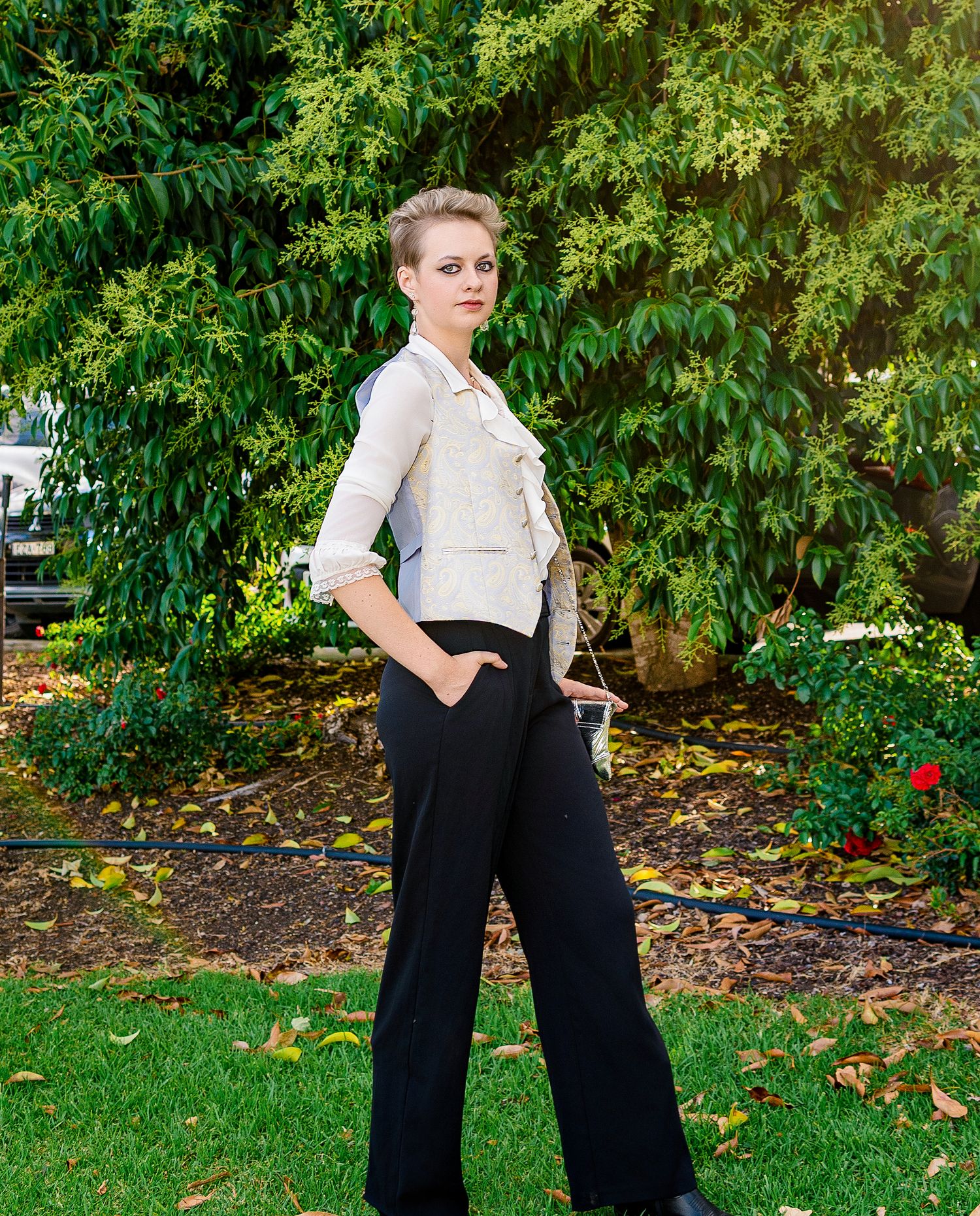 A woman with short hair poses outdoors in black wide-leg pants and a white ruffled blouse against lush green trees.