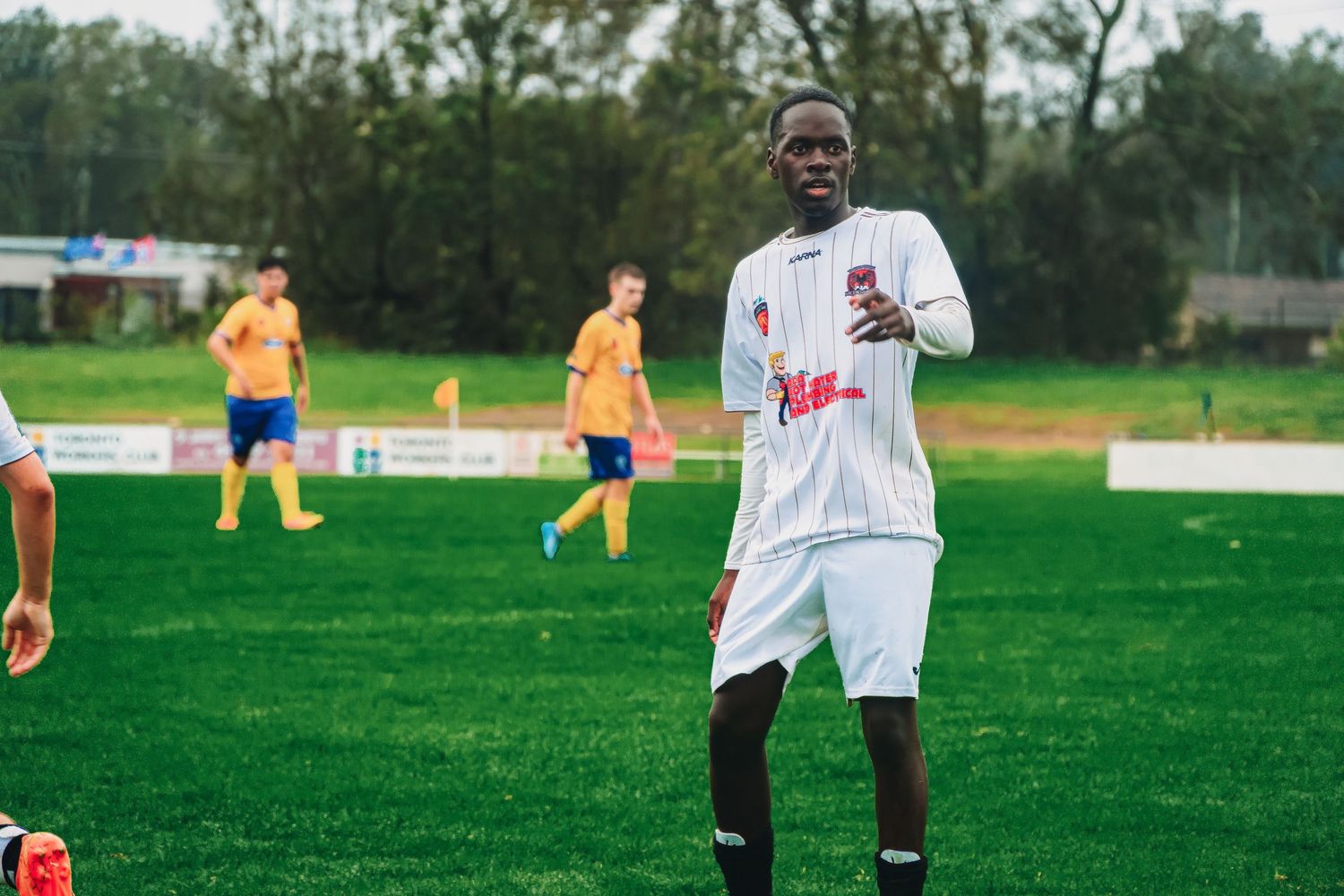 A soccer player in white jersey gestures on a green field during a match with teammates in yellow in the background.