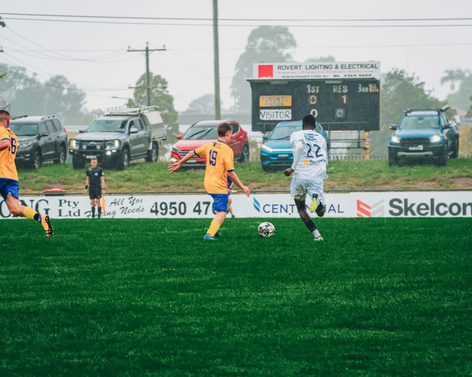 Soccer players in yellow and white jerseys compete on a green field during a misty outdoor match with scoreboards visible.