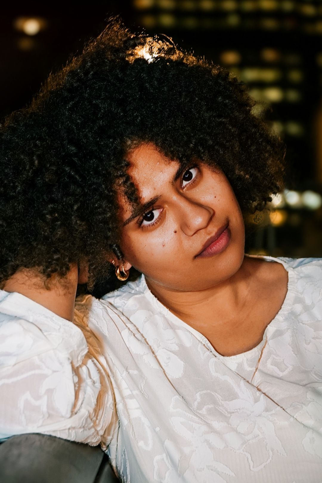Close-up portrait of curly-haired woman in white top with hands behind head at night.