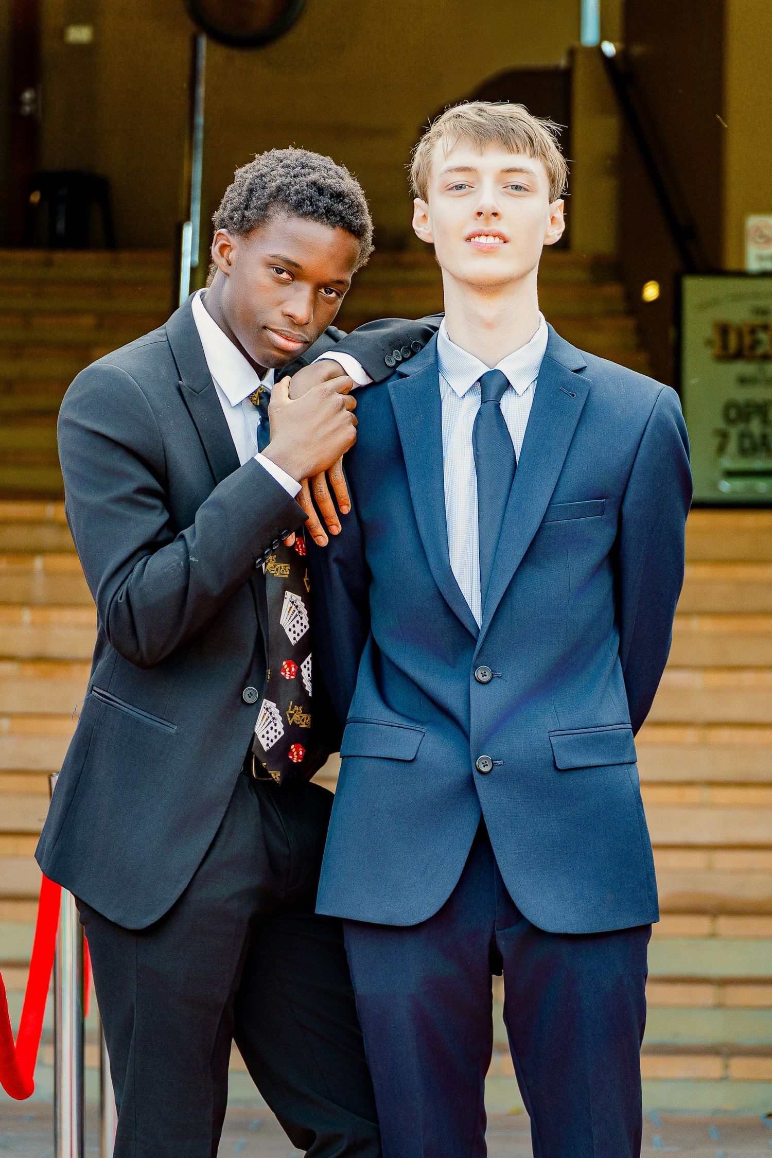 Two young men in formal suits pose together on steps at a prom or formal event.