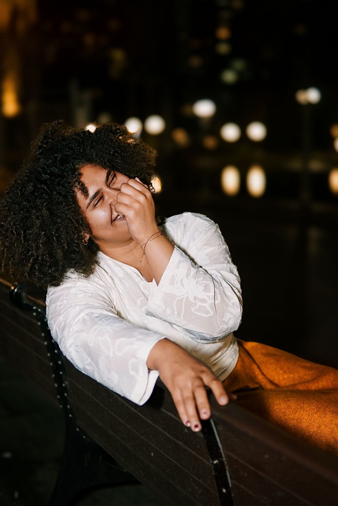 Woman with curly hair laughs joyfully while relaxing on a bench outdoors at night.