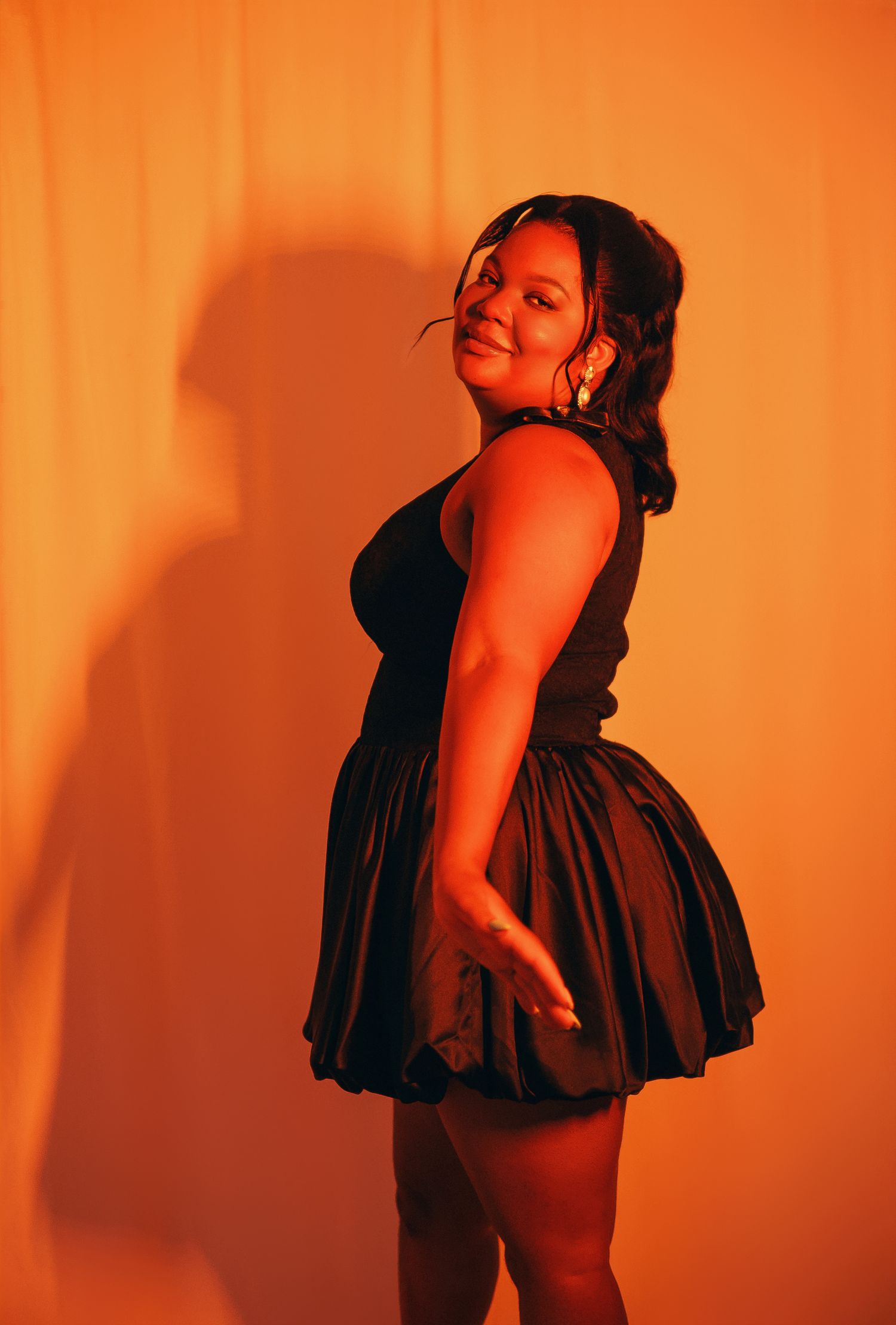 A curvy woman in a black dress poses confidently against an orange-lit backdrop, casting a dramatic shadow.