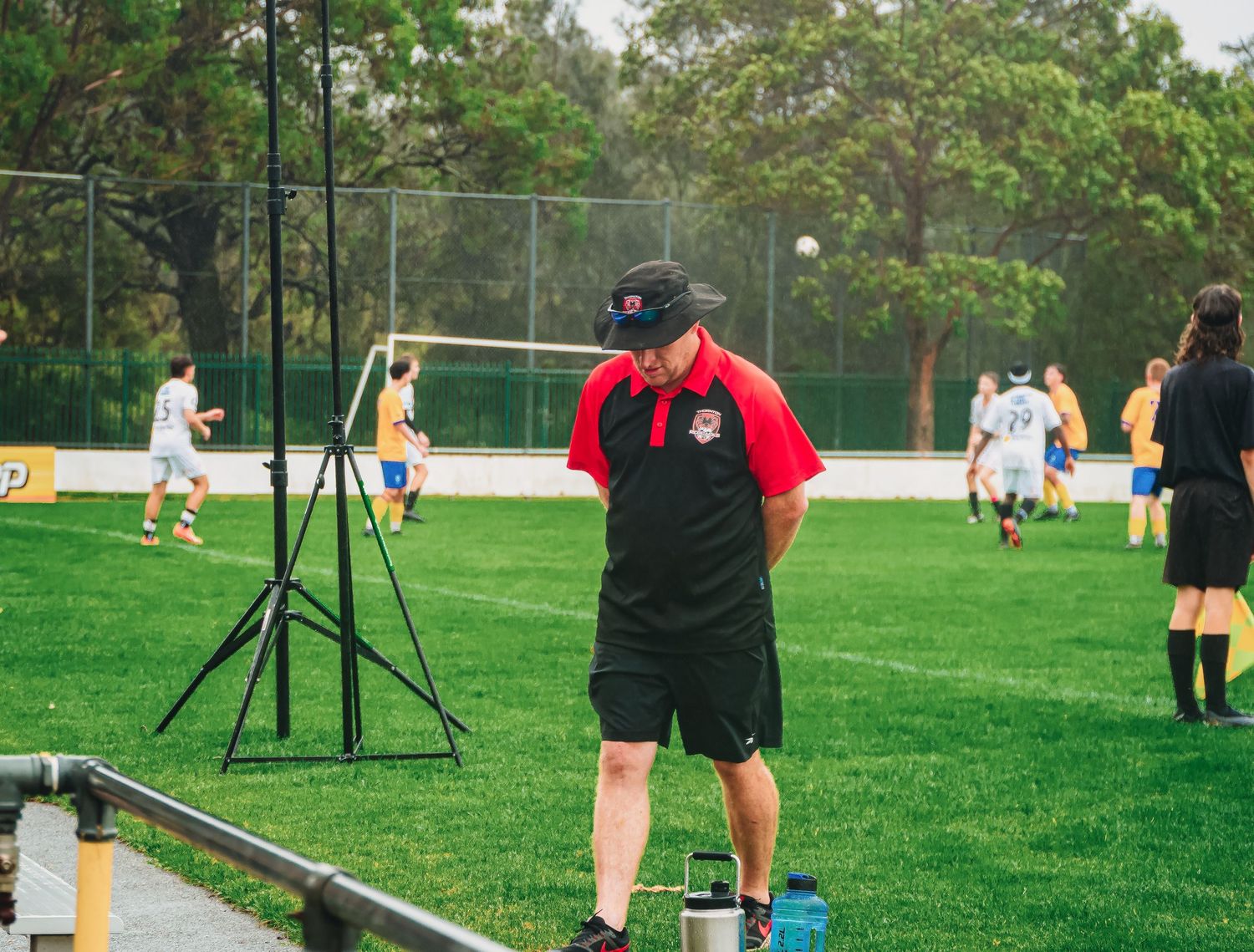 A soccer coach in red and black stands on the sideline observing a youth football match on a green field.