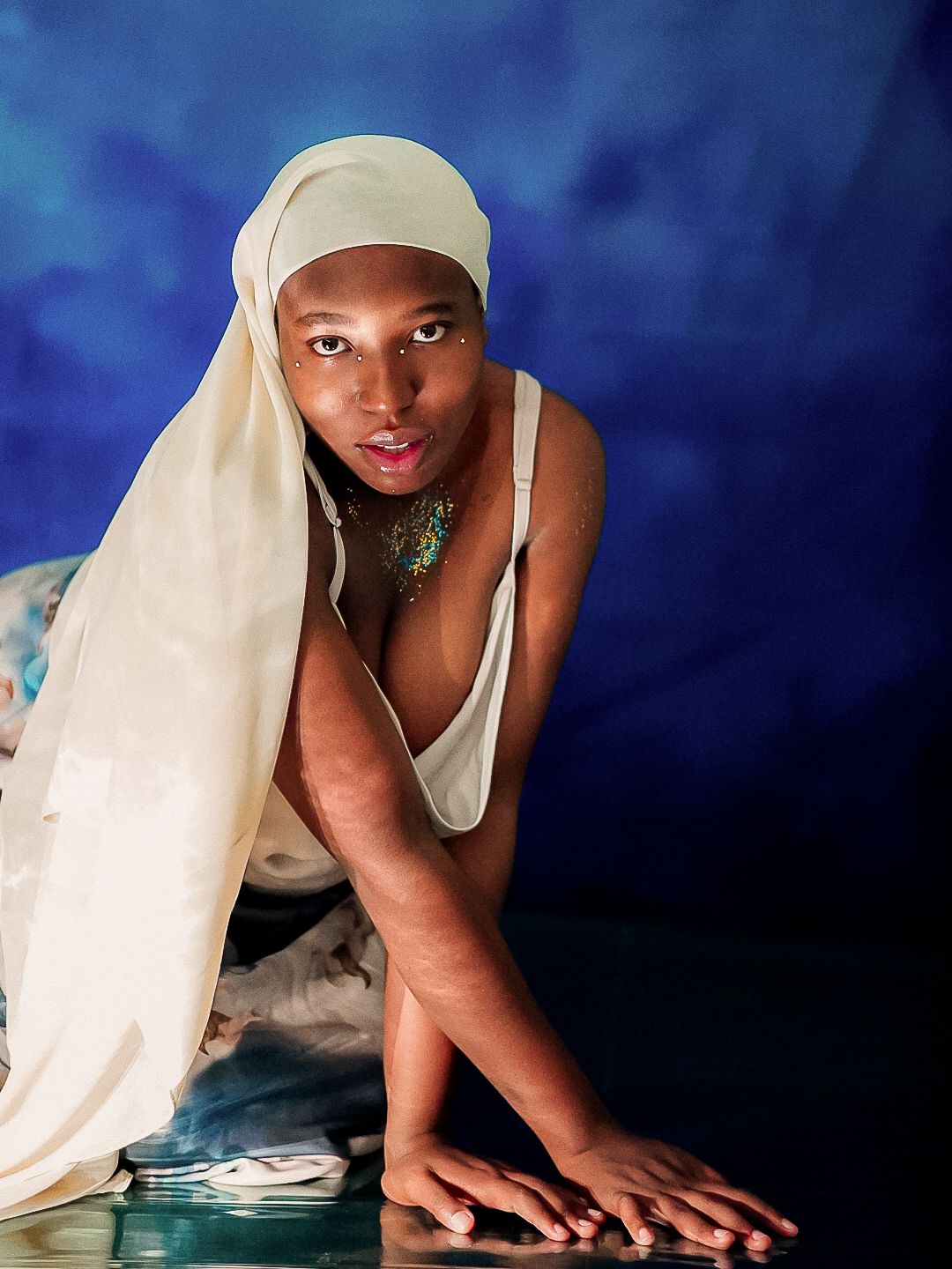 A young woman in a white headscarf and top poses gracefully on the floor against a blue background.