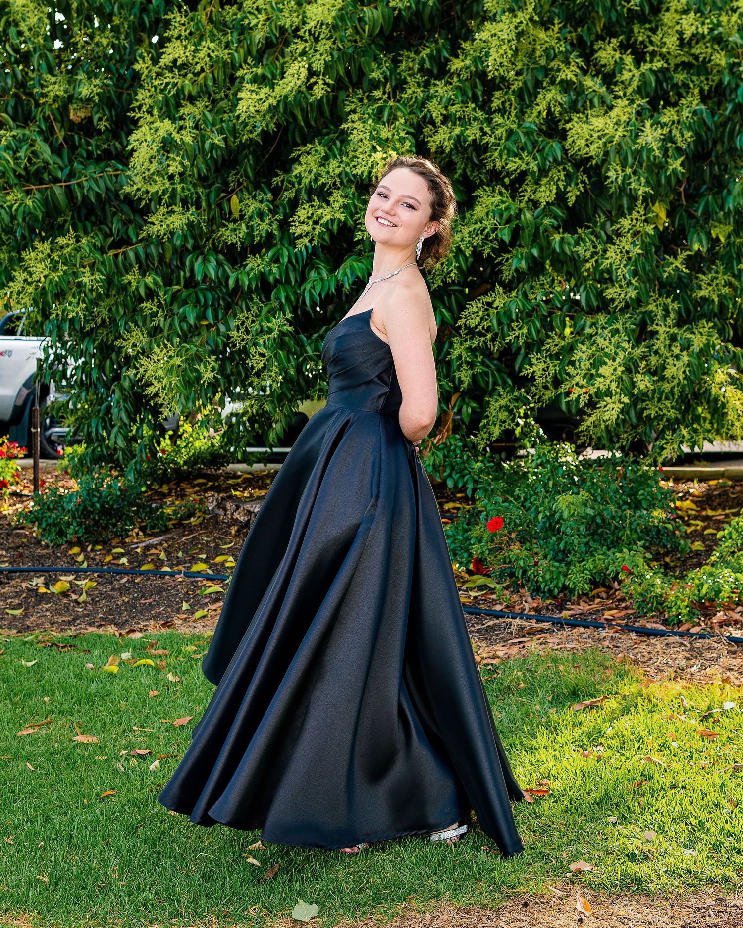 Young woman smiling in elegant black strapless ball gown outdoors in lush green garden setting.