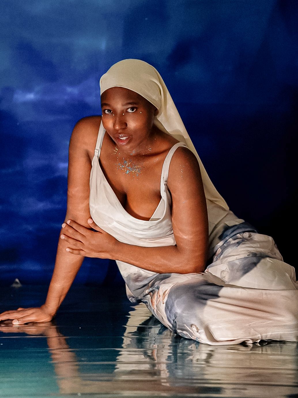 A woman in white draped fabric and headscarf poses gracefully near reflective water against a deep blue backdrop.