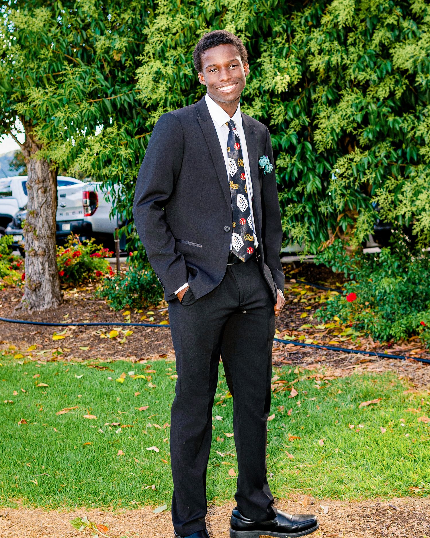 Young man in black suit with floral tie smiling outdoors on green lawn with trees in background.