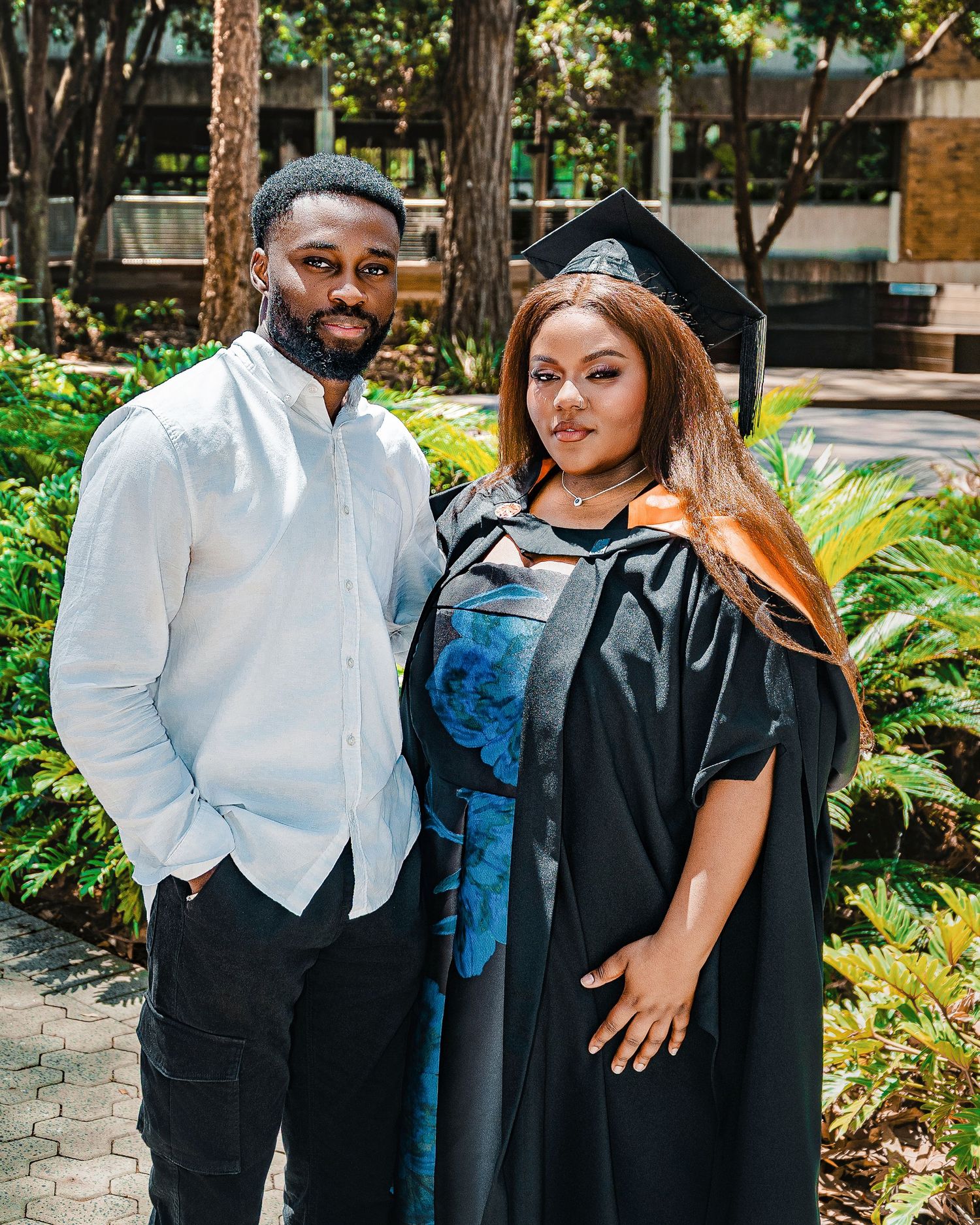 Graduate in cap and gown poses with a man in white shirt in a lush garden.