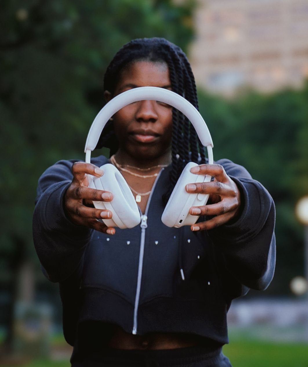 Young woman holding white headphones toward camera outdoors.
