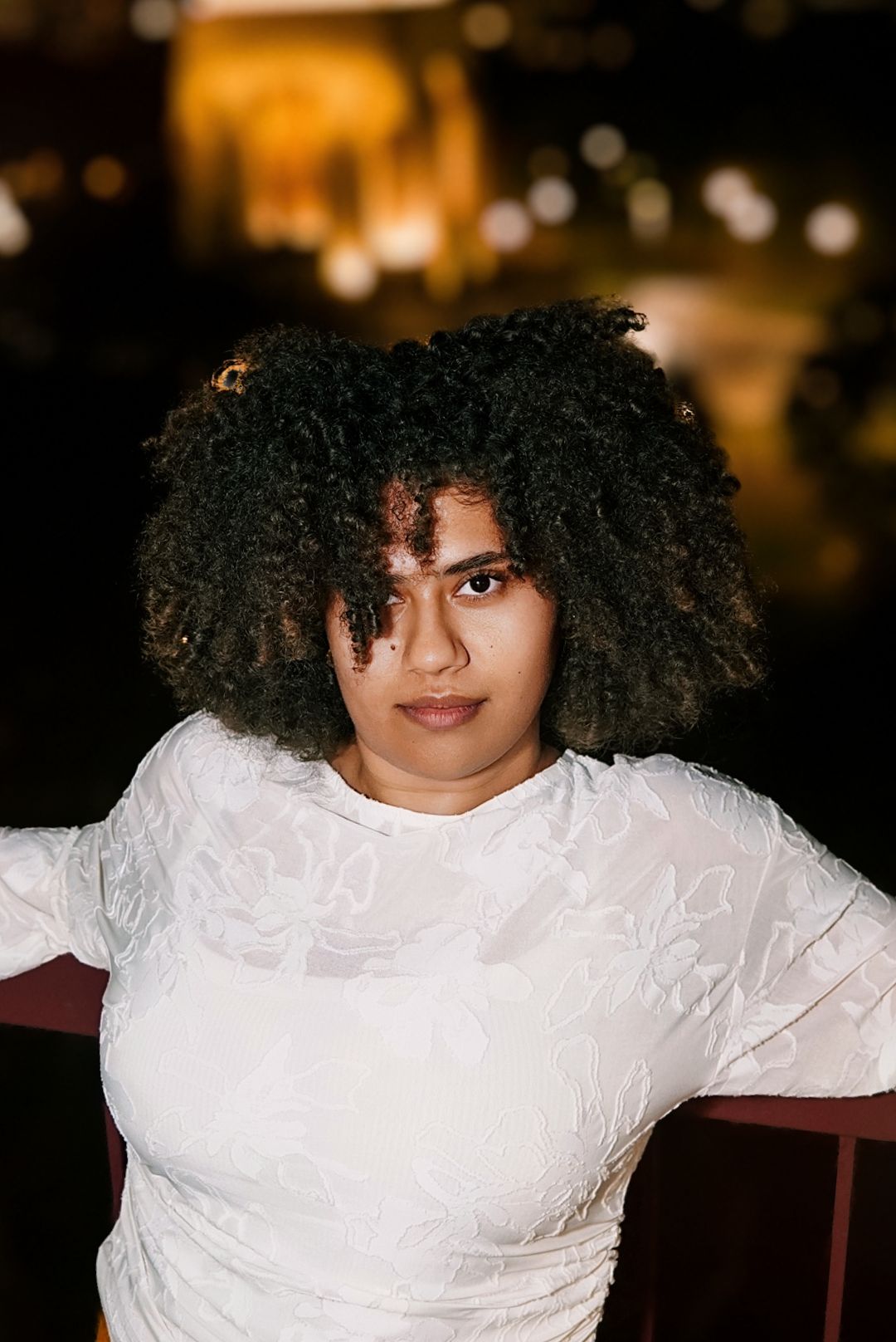 Close-up portrait of woman with natural curly hair in white lace top posing against night city backdrop.