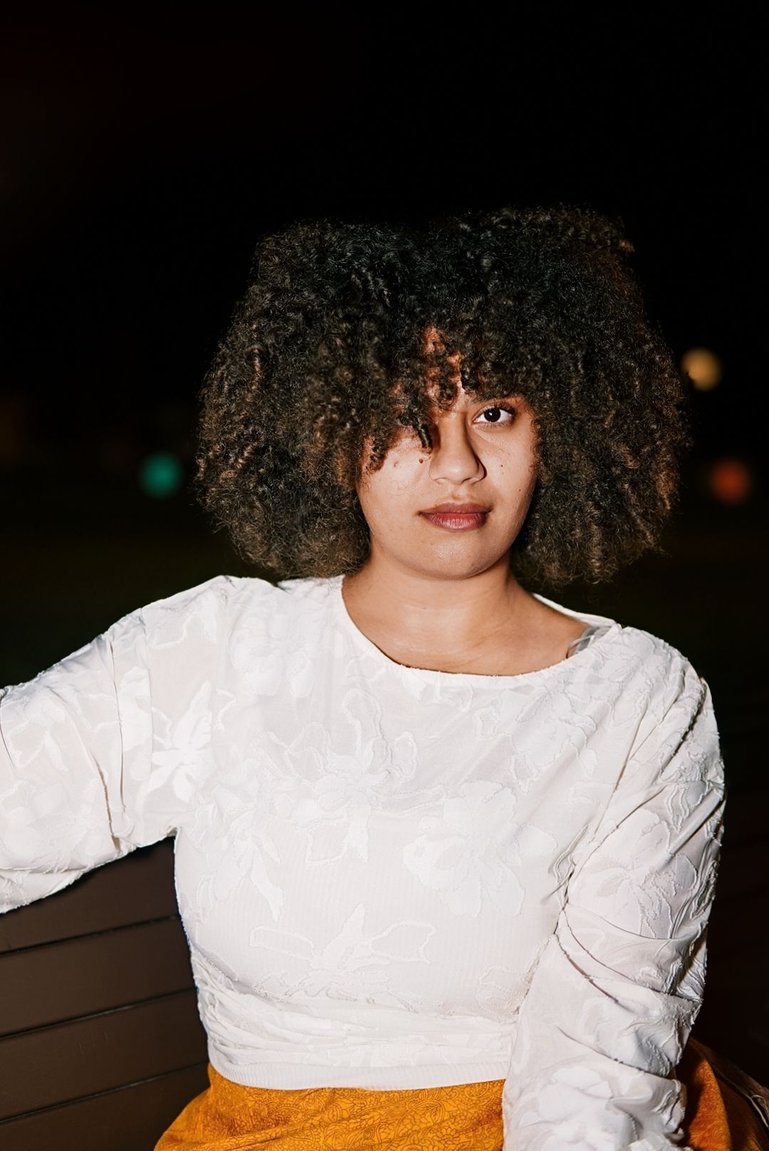 Young woman with curly afro hair wearing a white top and yellow skirt, sitting outdoors at night.